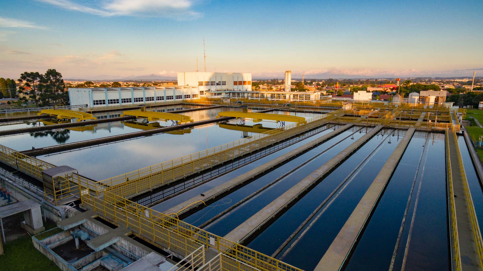 Vista aérea dos tanques de decantação e filtragem da ETA Iguaçu sob a luz do pôr do sol, com prédio operacional ao fundo.