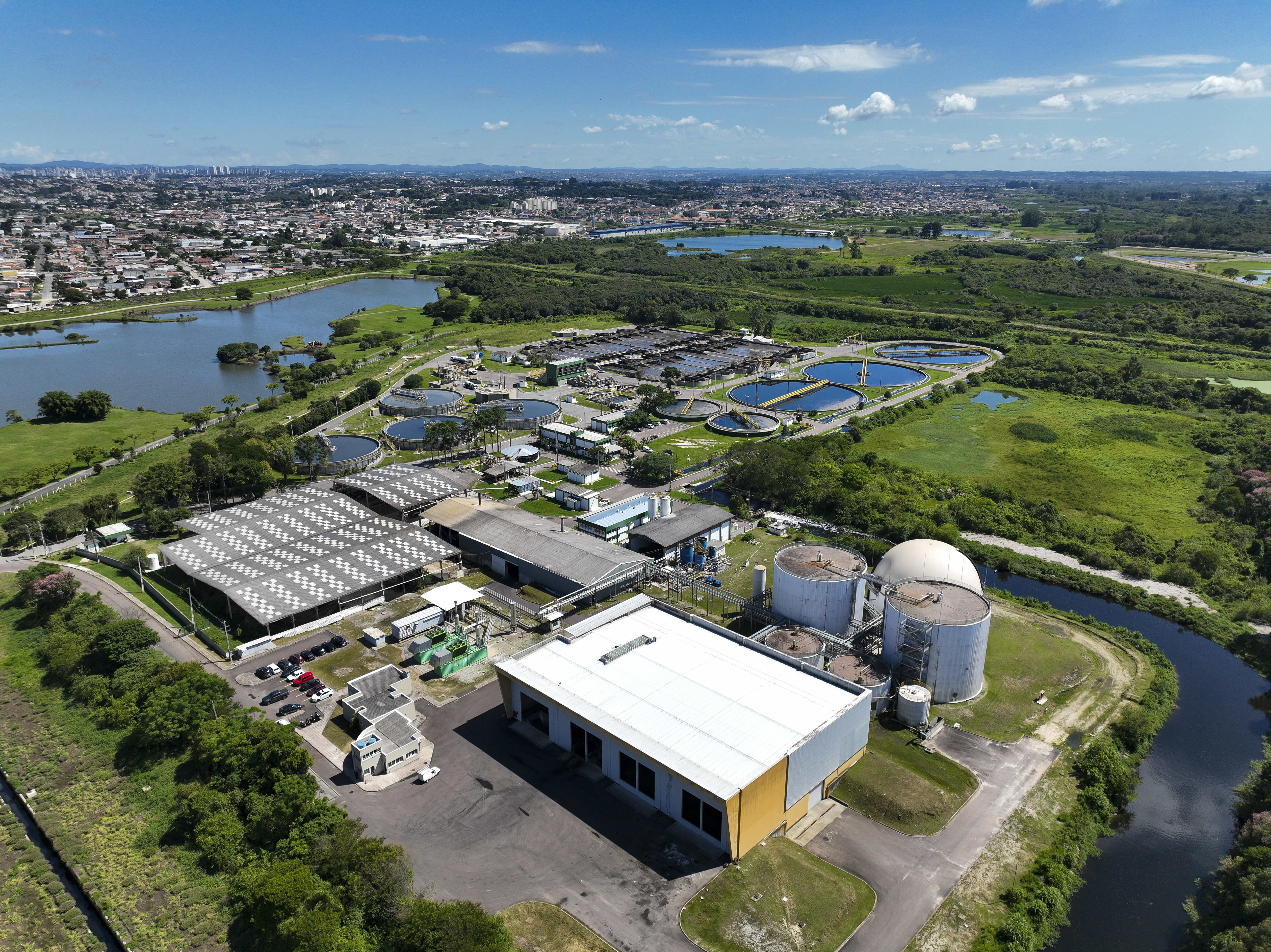 Vista aérea panorâmica da Estação de Tratamento de Esgoto Belém, exibindo os tanques circulares de decantação, lagoas de tratamento e os grandes biodigestores cilíndricos brancos ao lado do rio.