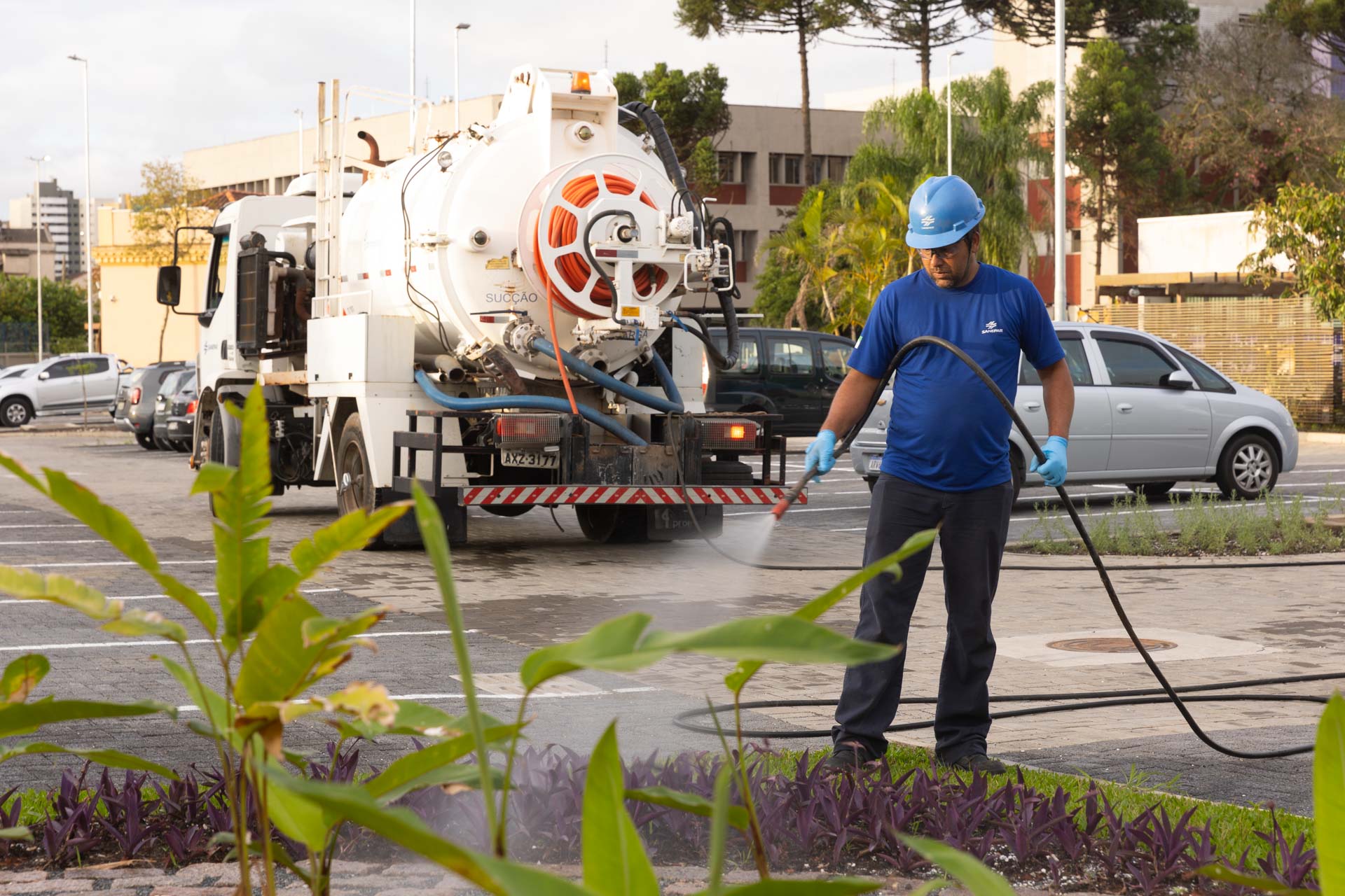 Um empregado da Sanepar (Companhia de Saneamento do Paraná), vestido com uniforme azul, capacete e luvas de proteção, utiliza uma mangueira conectada a um caminhão-tanque branco para regar um canteiro. A operação está sendo realizada com água de reúso.
