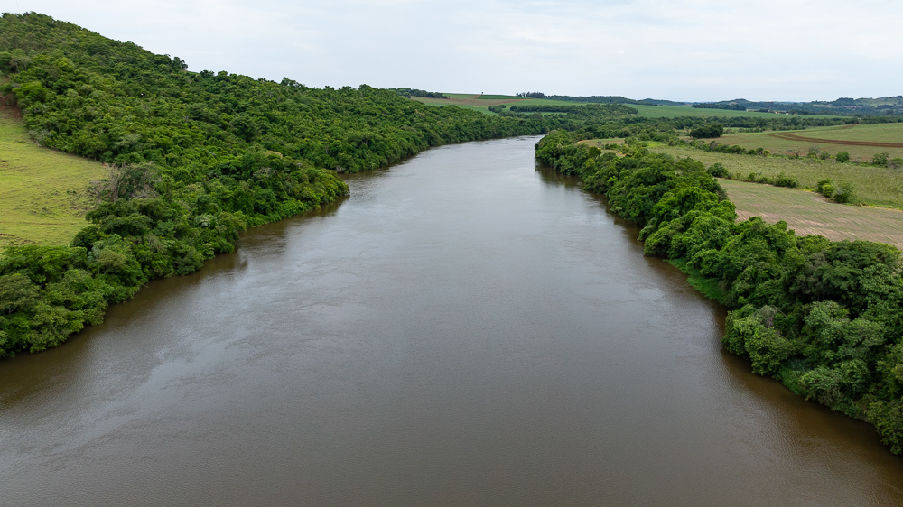 Vista aérea de um rio largo com águas de cor marrom turva, cercado por densa vegetação verde (mata ciliar) em ambas as margens. Ao fundo, paisagem com colinas e campos agrícolas sob um céu nublado.