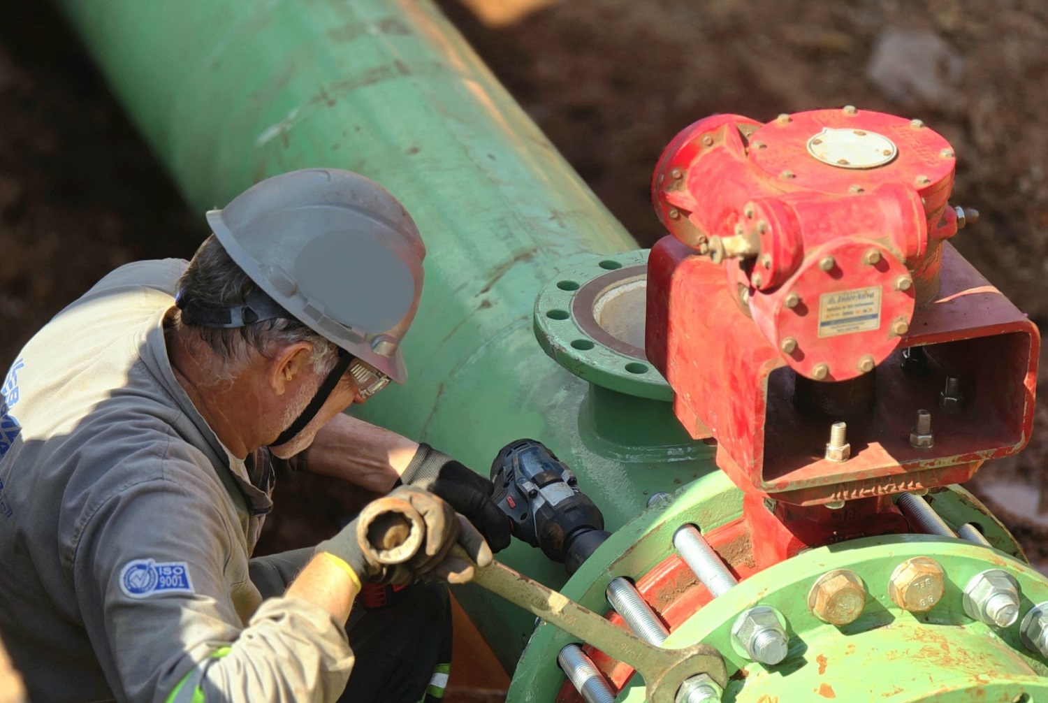 operário, devidamente equipado e com ferramentas, aperta parafusos em  uma conexão de tubulação 