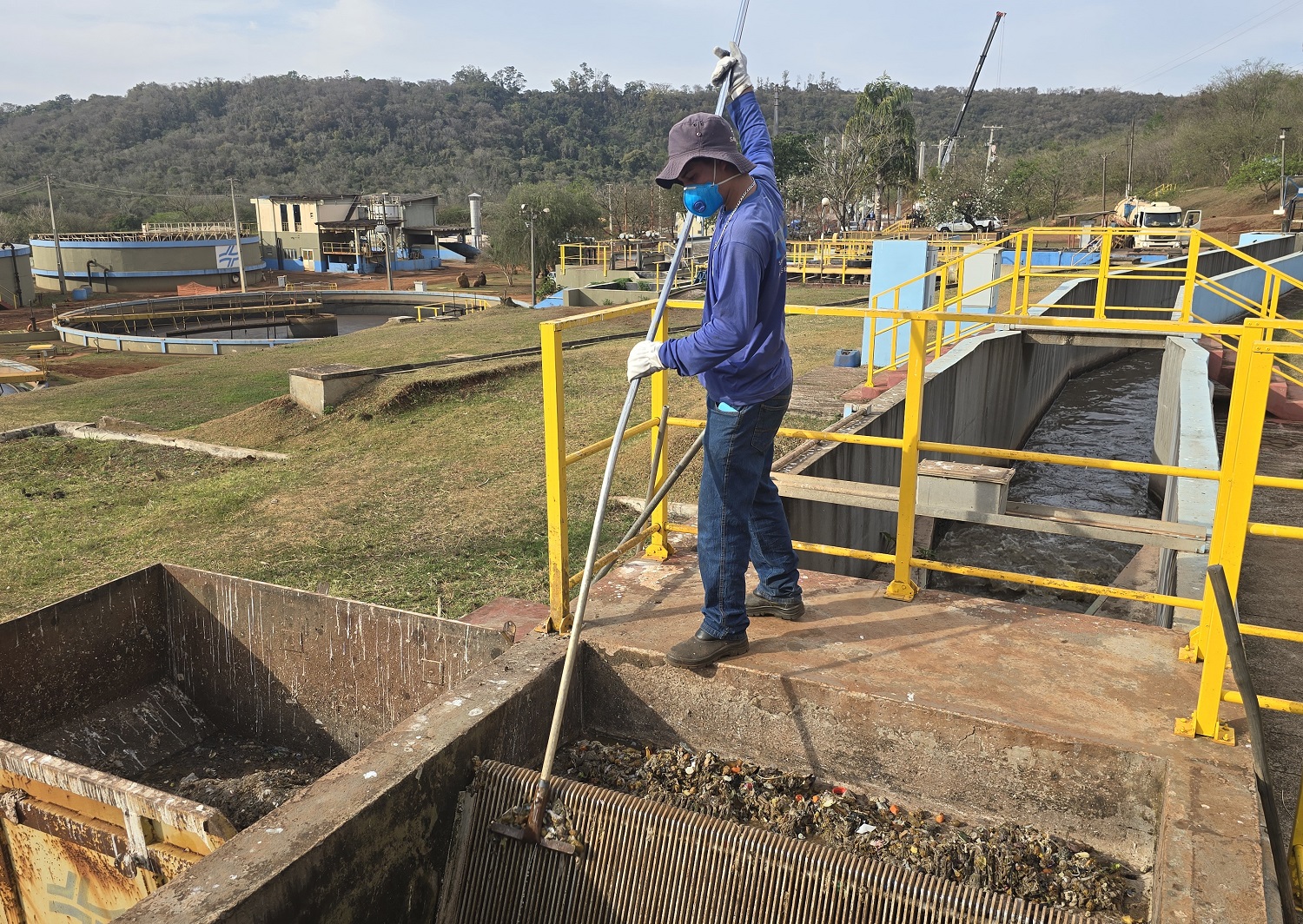 trabalhador devidamente equipado com máscara, retira resíduos sólidos de gradeamento na entrada da estação de tratamento de esgoto