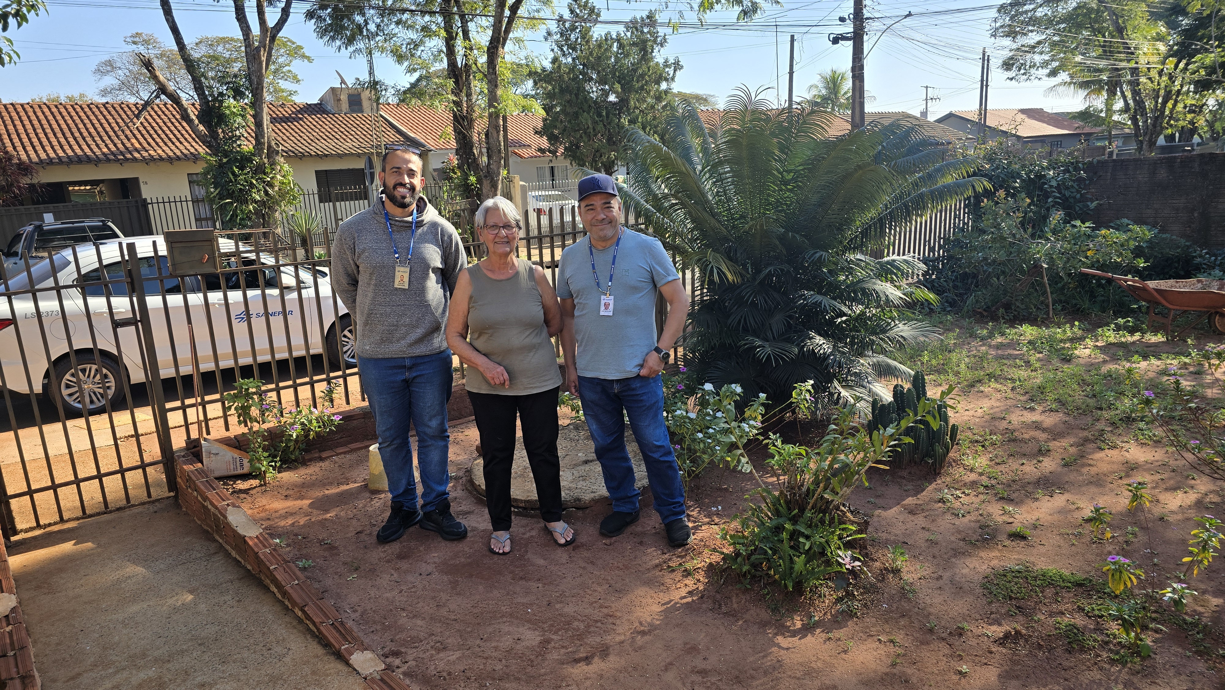 uma senhor de cabelos brancos está no quintal da sua casa, entre dois funcionários da sanepar 