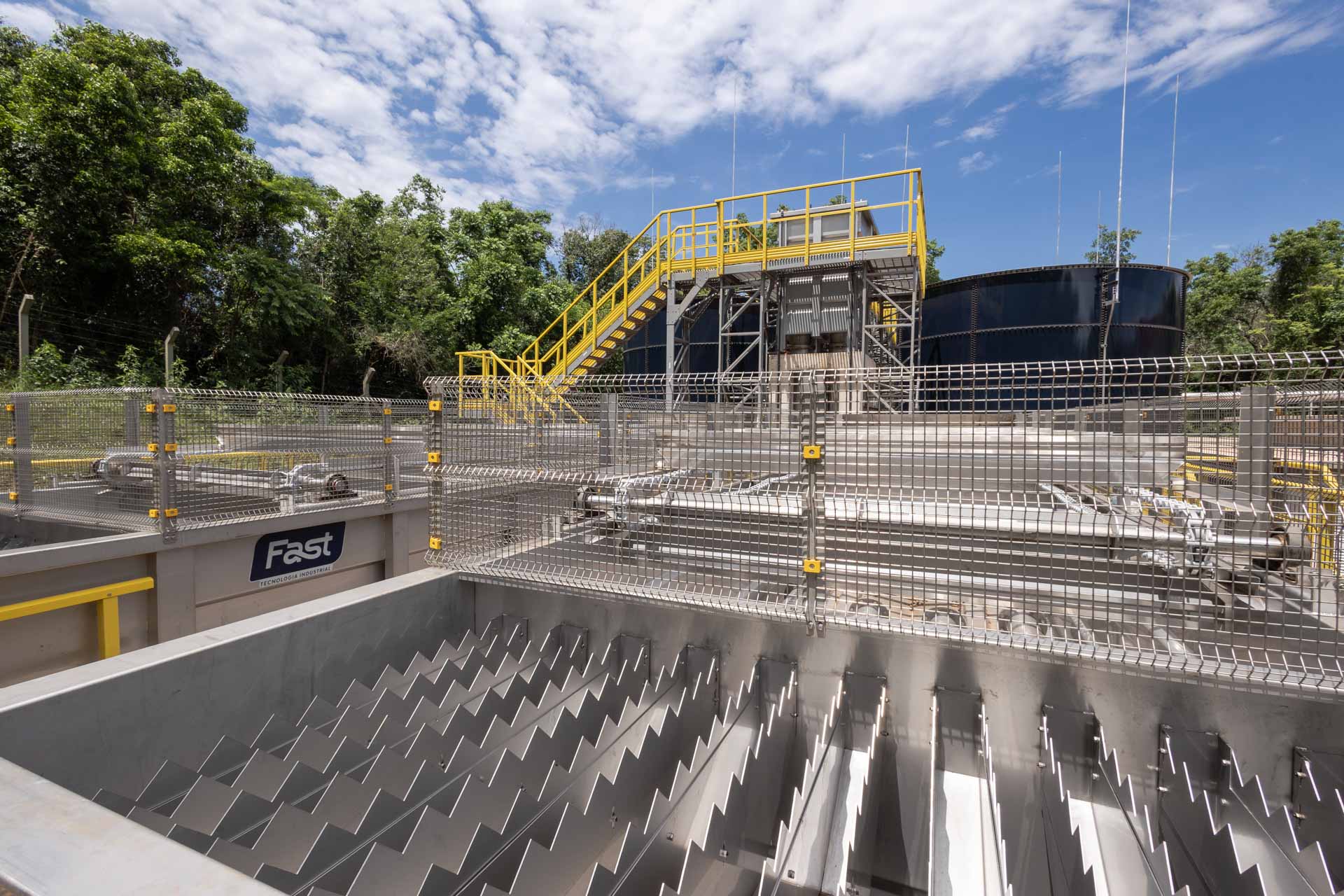 Estrutura metálica de tratamento de esgoto na ETE Alegria, em Medianeira, com grades de aço inoxidável em primeiro plano e uma escadaria amarela ao fundo sob céu azul