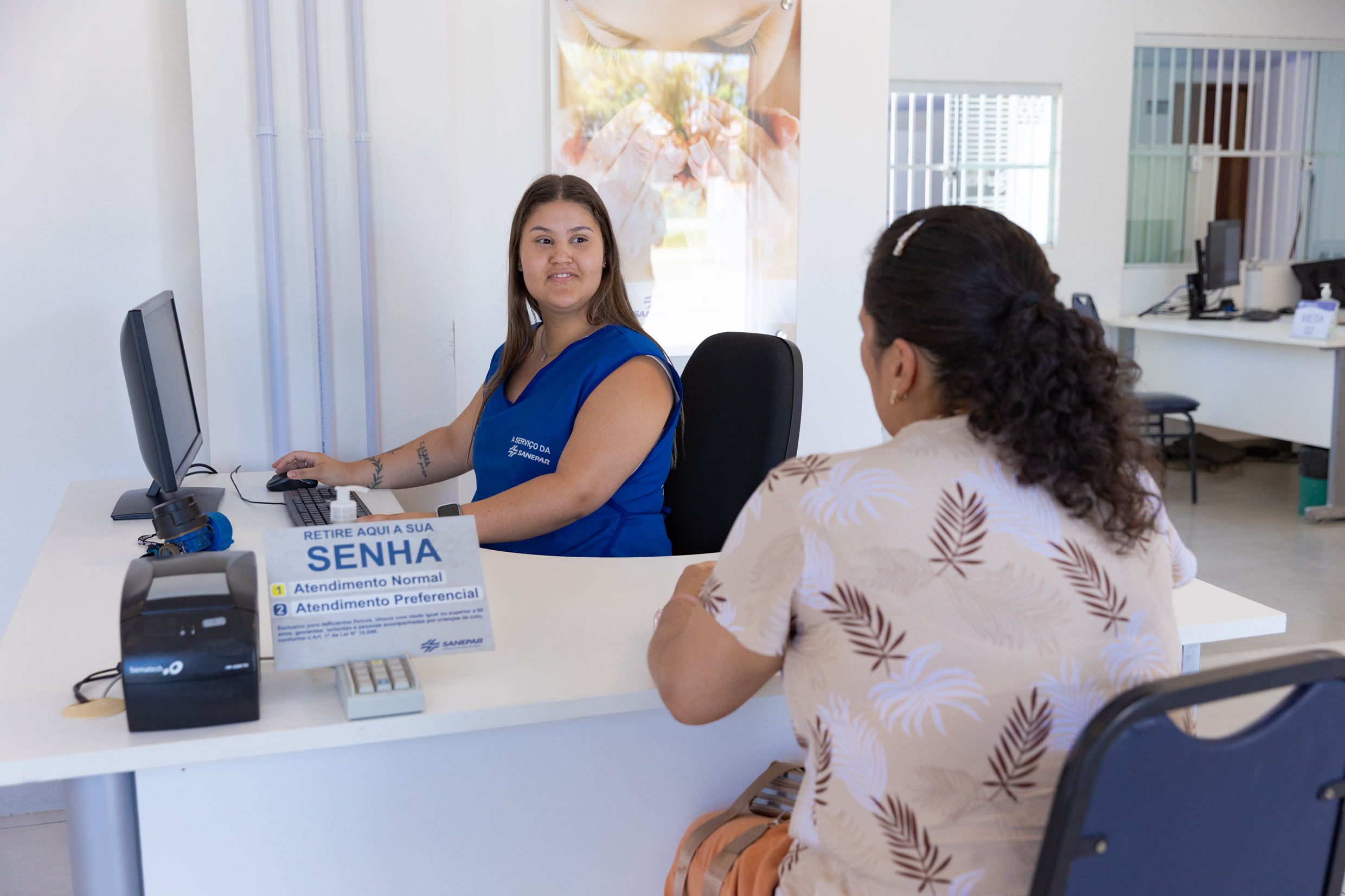 Atendente da Sanepar, vestindo colete azul da companhia, sorri ao prestar atendimento presencial para uma cliente sentada à sua frente. Sobre a mesa branca, há um computador e uma placa com a instrução para retirar senha de atendimento normal ou preferencial.