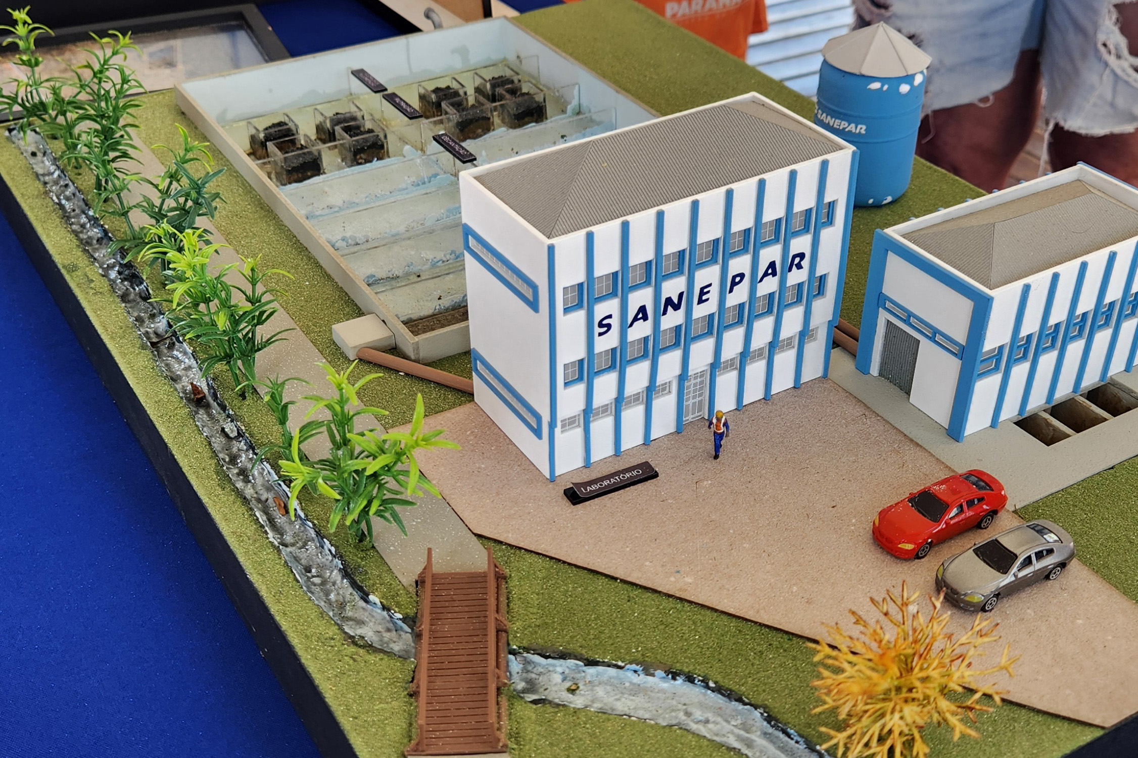 Maquete detalhada que simula uma Estação de Tratamento de Água (ETA) da Sanepar, com prédios administrativos, tanques de decantação, laboratório, reservatório e miniatura de carros.