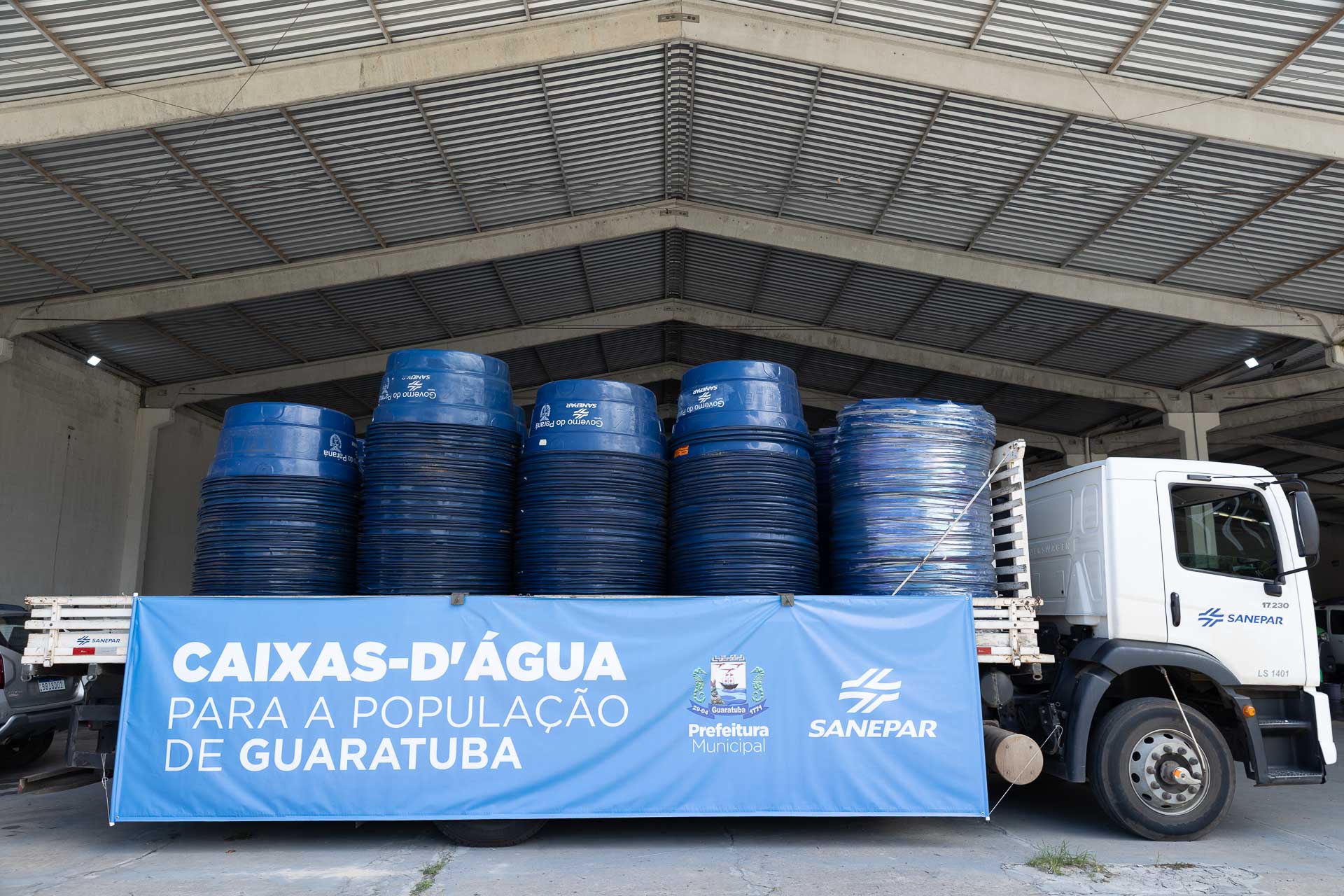 Um caminhão da Sanepar carregado de caixas-d'água azuis empilhadas está estacionado em um galpão. Uma faixa azul na lateral do caminhão tem o texto 'CAIXAS-D'ÁGUA PARA A POPULAÇÃO DE GUARATUBA' e os logotipos da Prefeitura Municipal e da Sanepar.