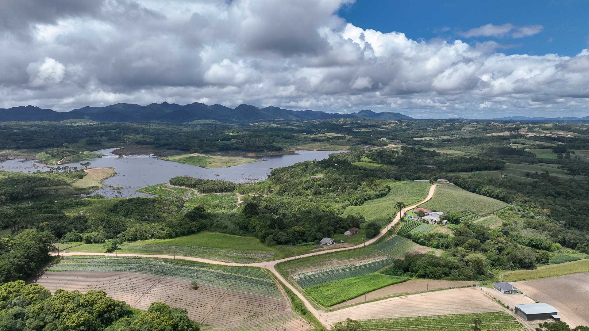Vista aérea de represa cercada por vegetação nativa e propriedades rurais com áreas agrícolas e estradas de terra, com serras no horizonte sob céu com nuvens.