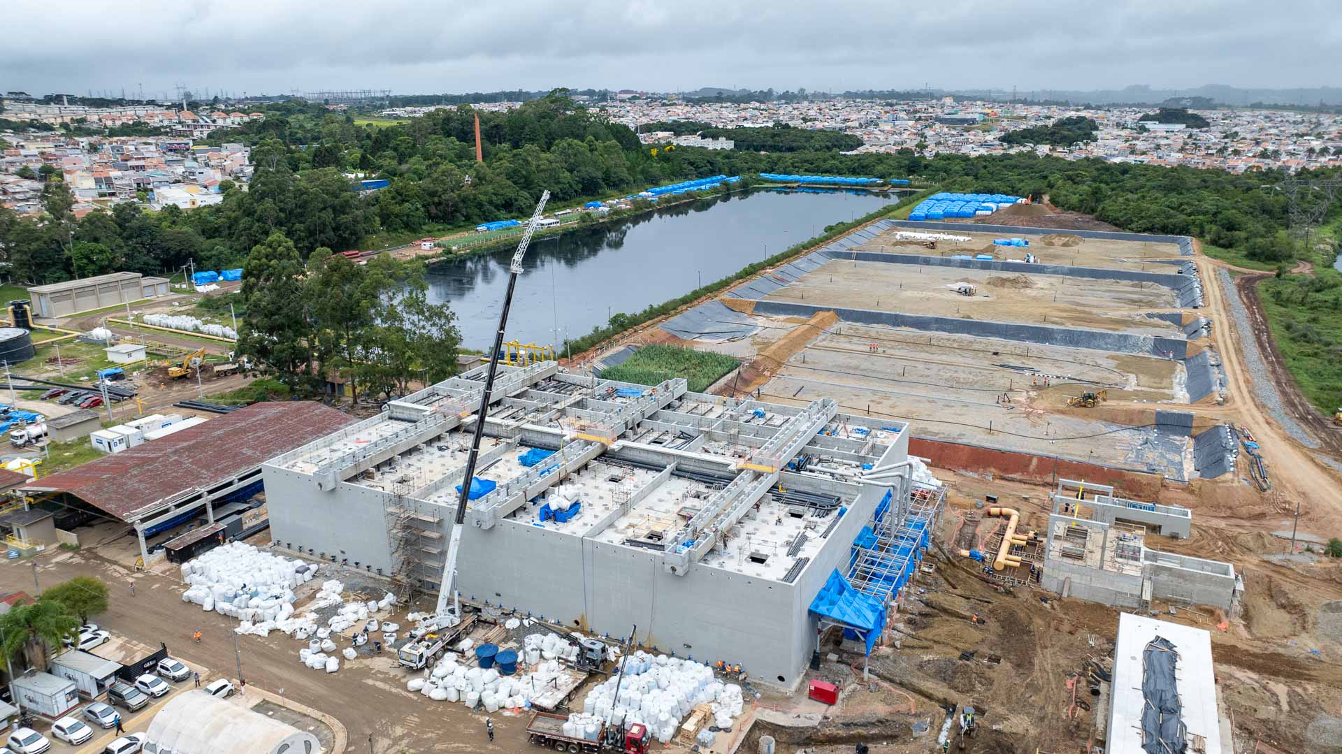 Vista aérea da ampla obra de construção da ETE CIC Xisto, com uma grande estrutura retangular de concreto em destaque, guindaste em operação, lagoa ao fundo e tanques de tratamento biológico por Wetland em andamento
