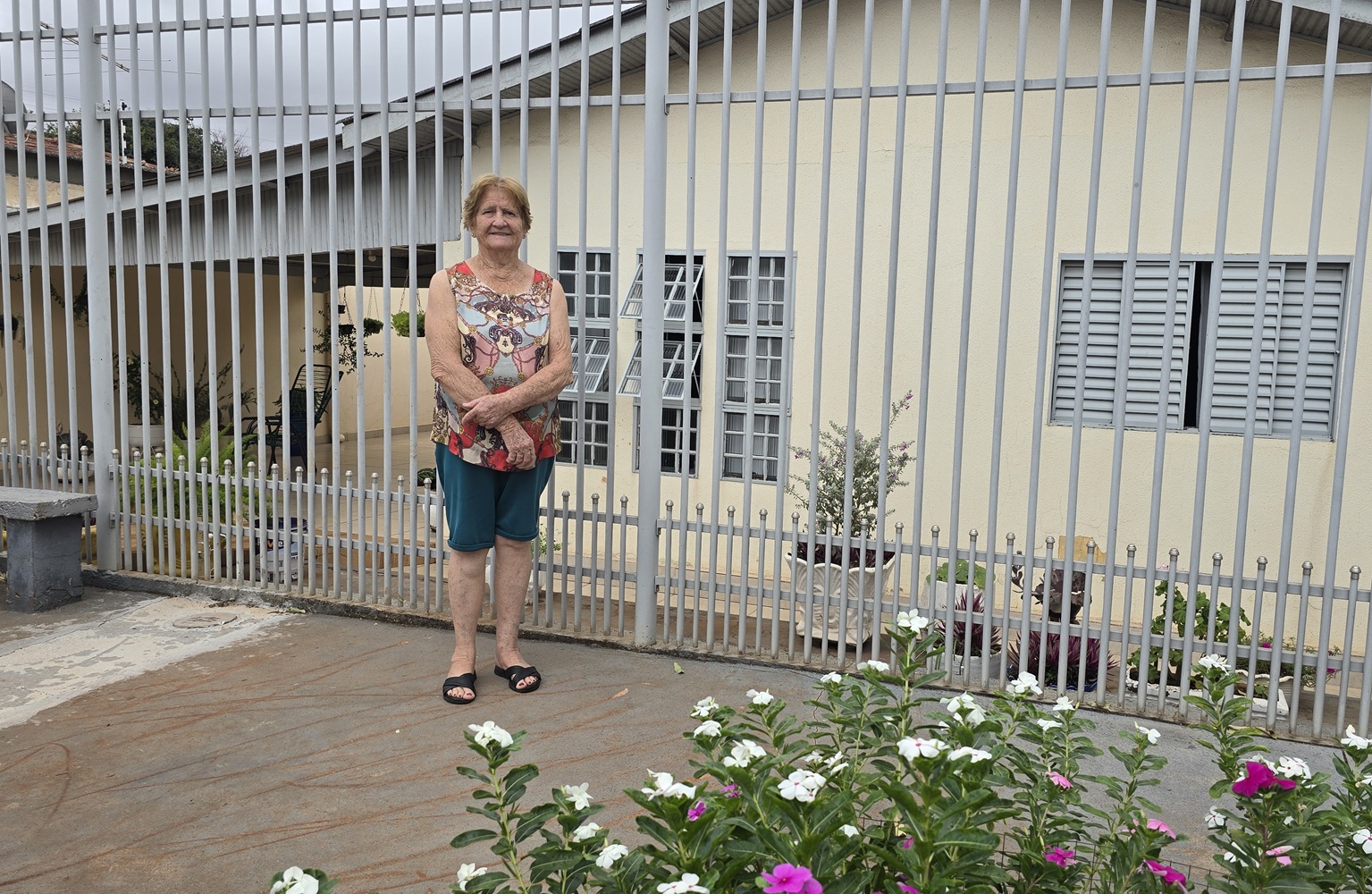 uma senhora loira de cabelos curtos está na frente da sua casa, ao fundo o quintal com flores e a fossa no chão