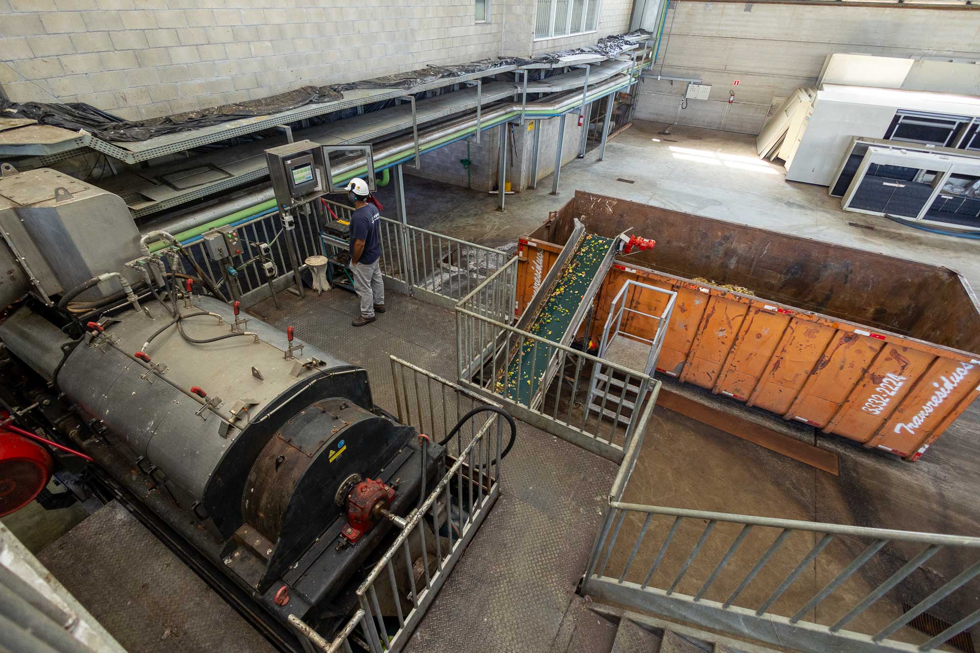 Funcionário opera painel eletrônico de controle em área industrial, ao lado de maquinário mecânico e de uma esteira transportadora que descarta resíduos de alimentos em uma caçamba laranja.
