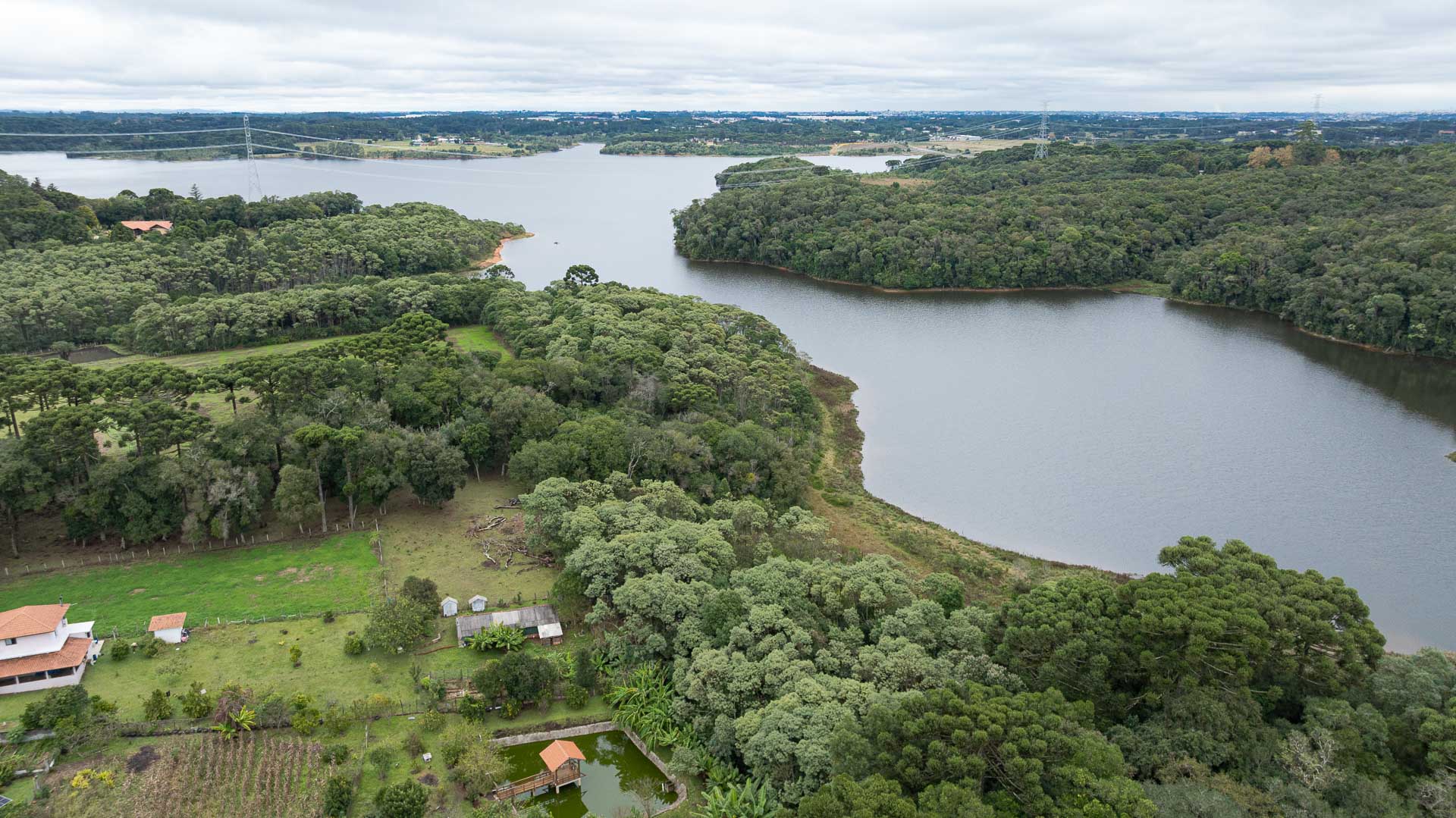 Vista aérea ampla que mostra a sinuosidade de uma represa circundada pela Mata Atlântica. O corpo d'água azul-escuro serpenteia da direita para o centro da imagem, estendendo-se até o horizonte. No canto inferior esquerdo, há uma propriedade rural com um gramado verde-claro bem cuidado, uma casa de dois andares com telhado de cerâmica avermelhada, um pequeno açude retangular com um trapiche de madeira cobertinho e pequenas hortas. A floresta densa, rica em araucárias, abraça toda a margem da represa.