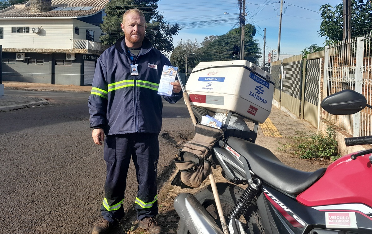 A imagem mostra o mesmo trabalhador da foto anterior, desta vez de corpo inteiro, em pé na rua ao lado de uma moto. Ele veste o mesmo uniforme azul-escuro com faixas reflexivas e o crachá da Sanepar e Enorsul