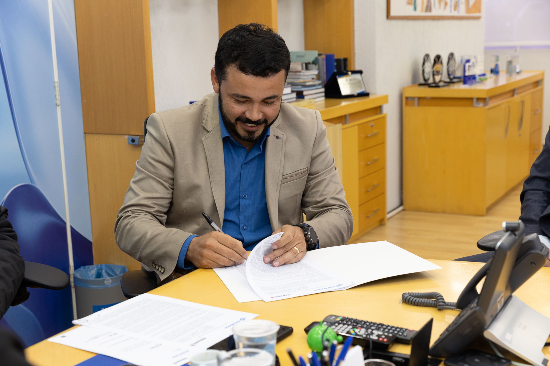 Em primeiro plano, o prefeito de Guaraqueçaba, vestindo paletó claro sobre camisa social azul, aparece sentado à mesa amarela assinando papéis. Ele está sorrindo e concentrado na assinatura.