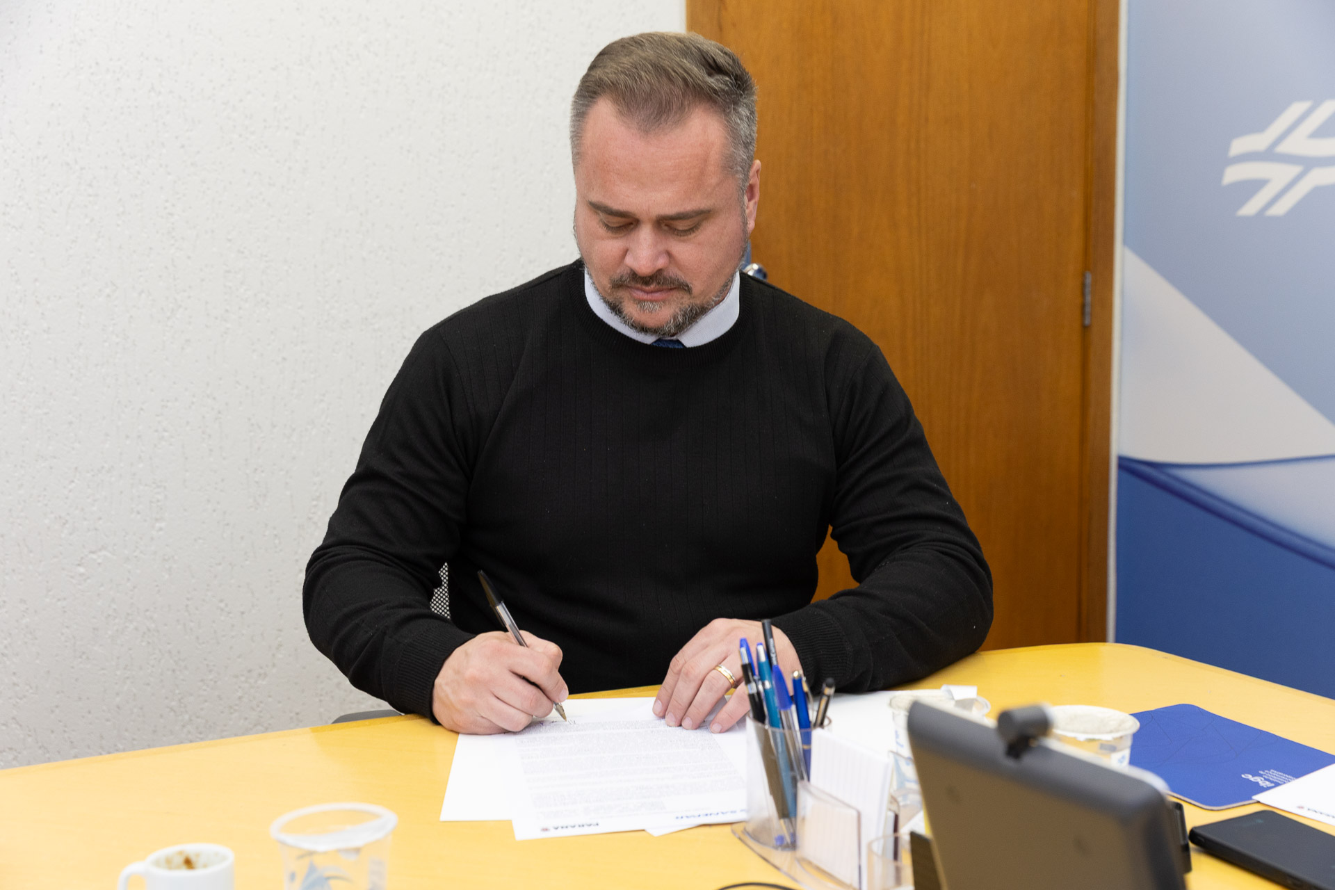 Em primeiro plano, o deputado estadual Alisson Wandscheer, vestindo blusa preta de gola alta sobre camisa clara, aparece sentado à mesa amarela assinando um documento com caneta preta. Ele está concentrado, com o olhar voltado para o papel.