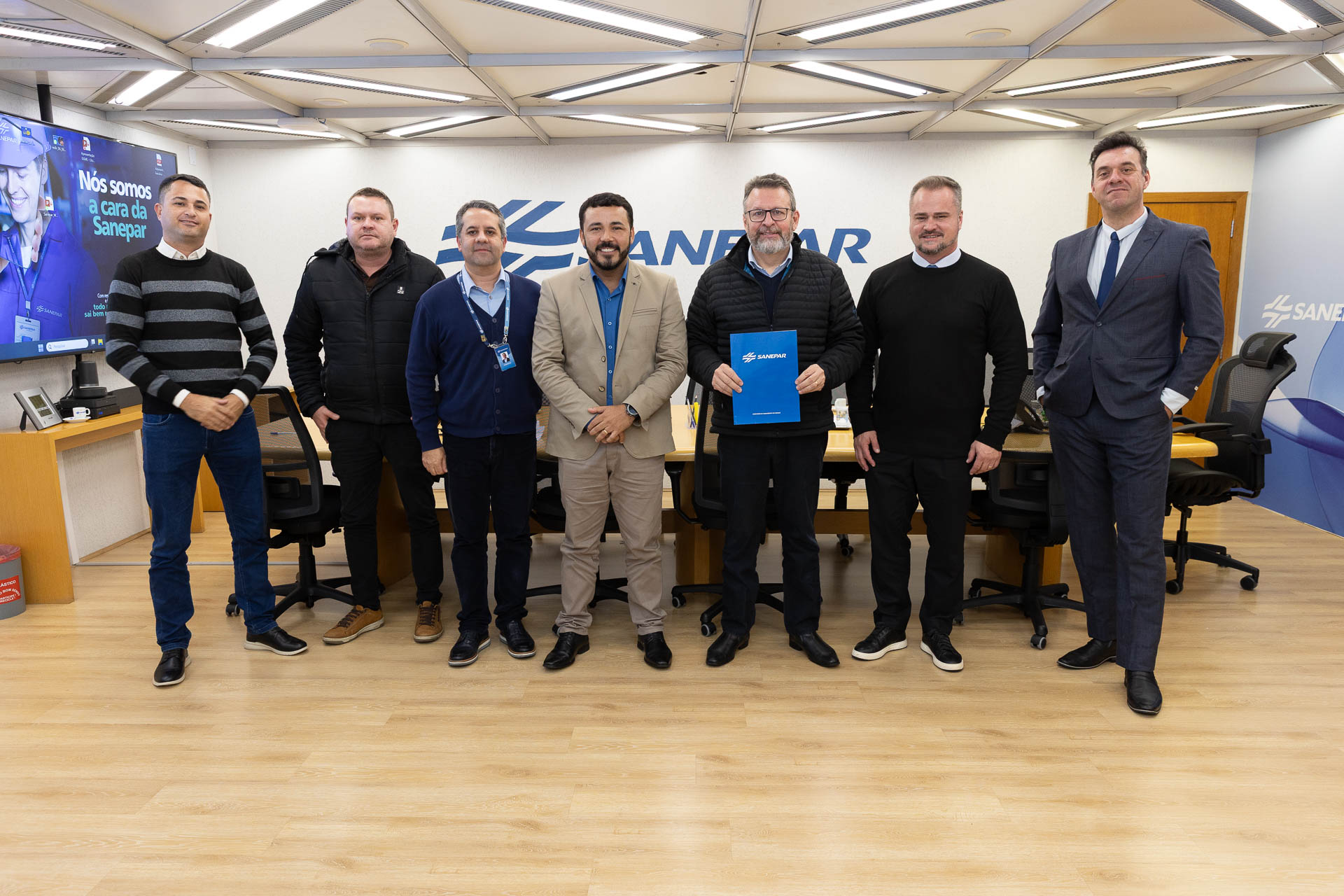 Sete homens alinhados lado a lado em uma sala de reuniões da Sanepar. No centro, o prefeito de Guaraqueçaba, Alessandro Carneiro Soares Truchinski, de paletó claro sorri e, ao seu lado, o diretor-presidente da Sanepar, Wilson Bley, segura uma pasta azul com a marca da Sanepar. Ao fundo, destaca-se o logotipo da companhia na parede.