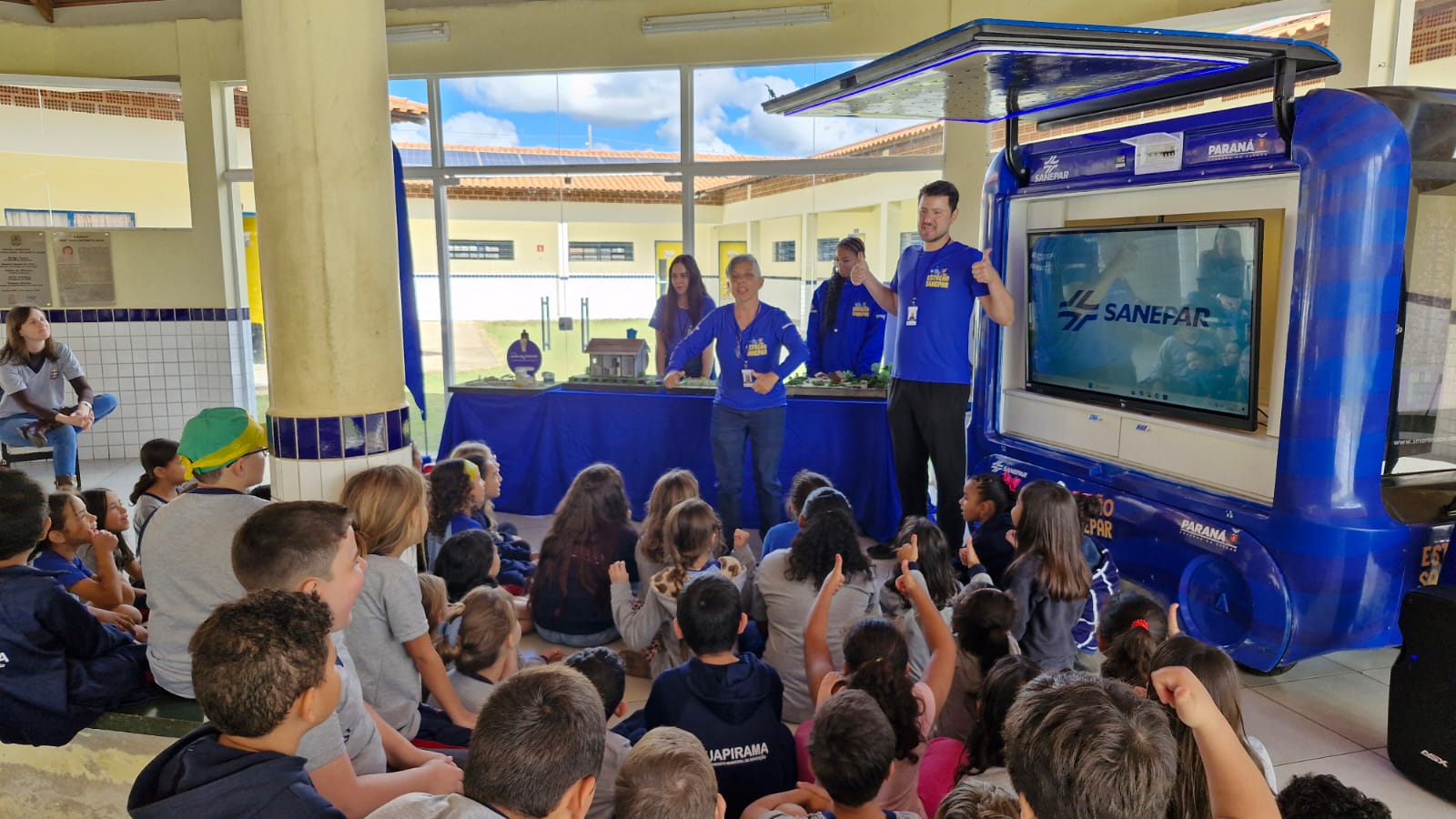 Monitores da Sanepar gesticulam alegremente durante uma palestra educativa sobre saneamento para crianças sentadas no pátio da Escola Maria Nagib, em Guapirama. Ao lado, há uma van adaptada com uma grande tela de TV.