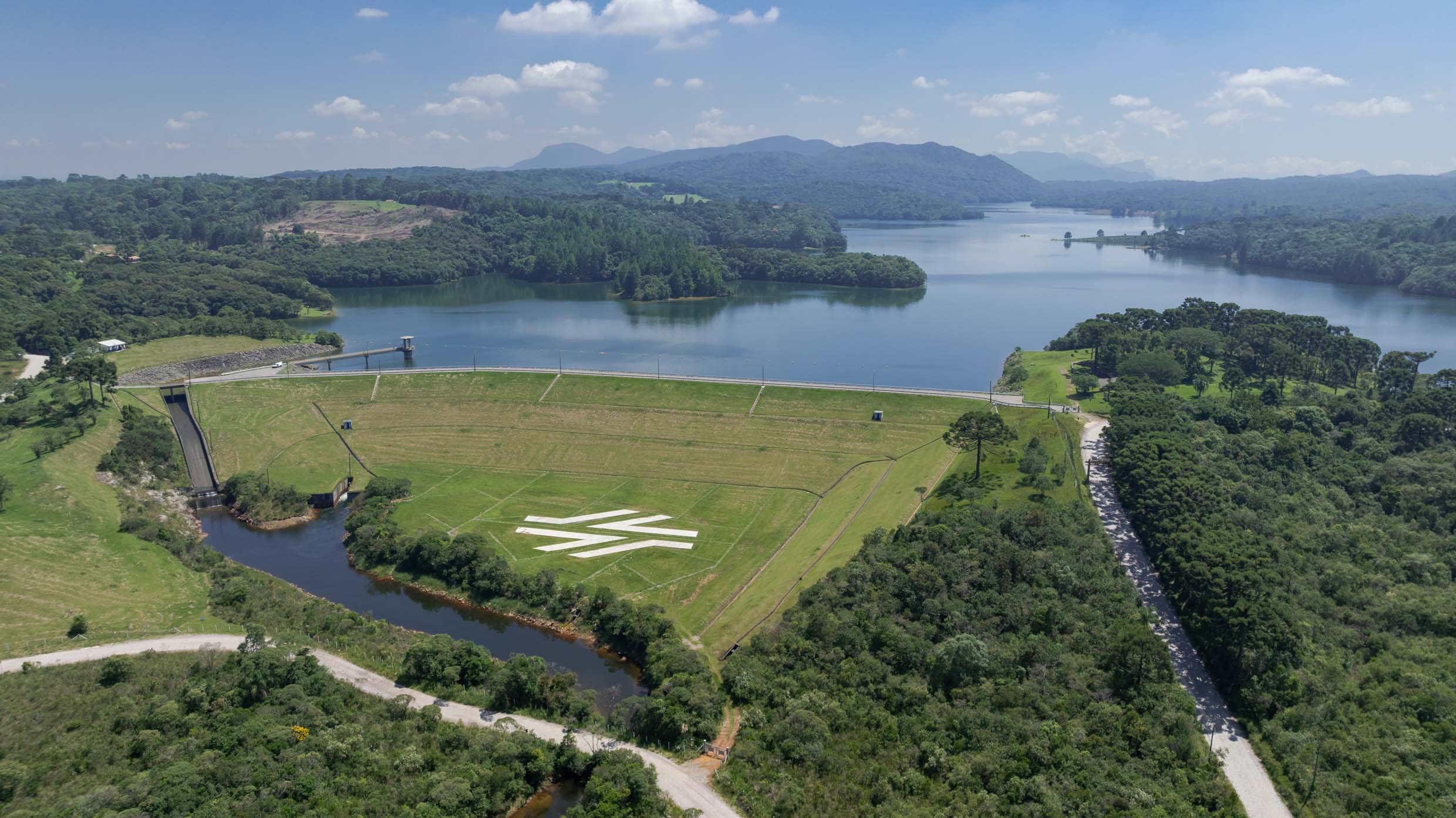 Vista aérea da Barragem de Piraquara com o reservatório de água ao fundo. No gramado do talude da barragem, destaca-se o logotipo estilizado da Sanepar em cor branca