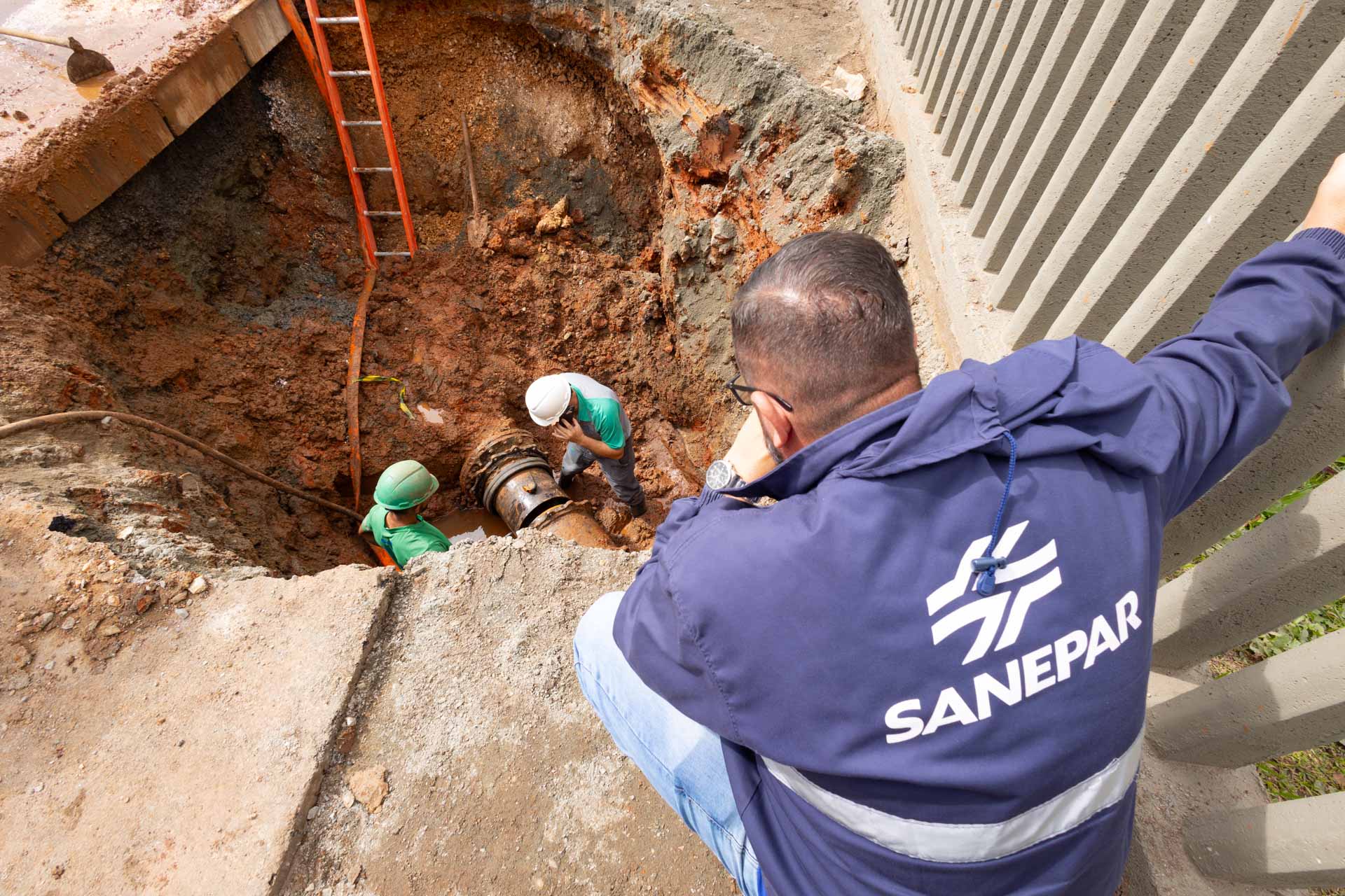 Equipe técnica trabalha na manutenção de uma grande tubulação de água dentro de uma escavação na terra. Em primeiro plano, um supervisor de costas, vestindo jaqueta azul escura com a marca Sanepar, observa o serviço