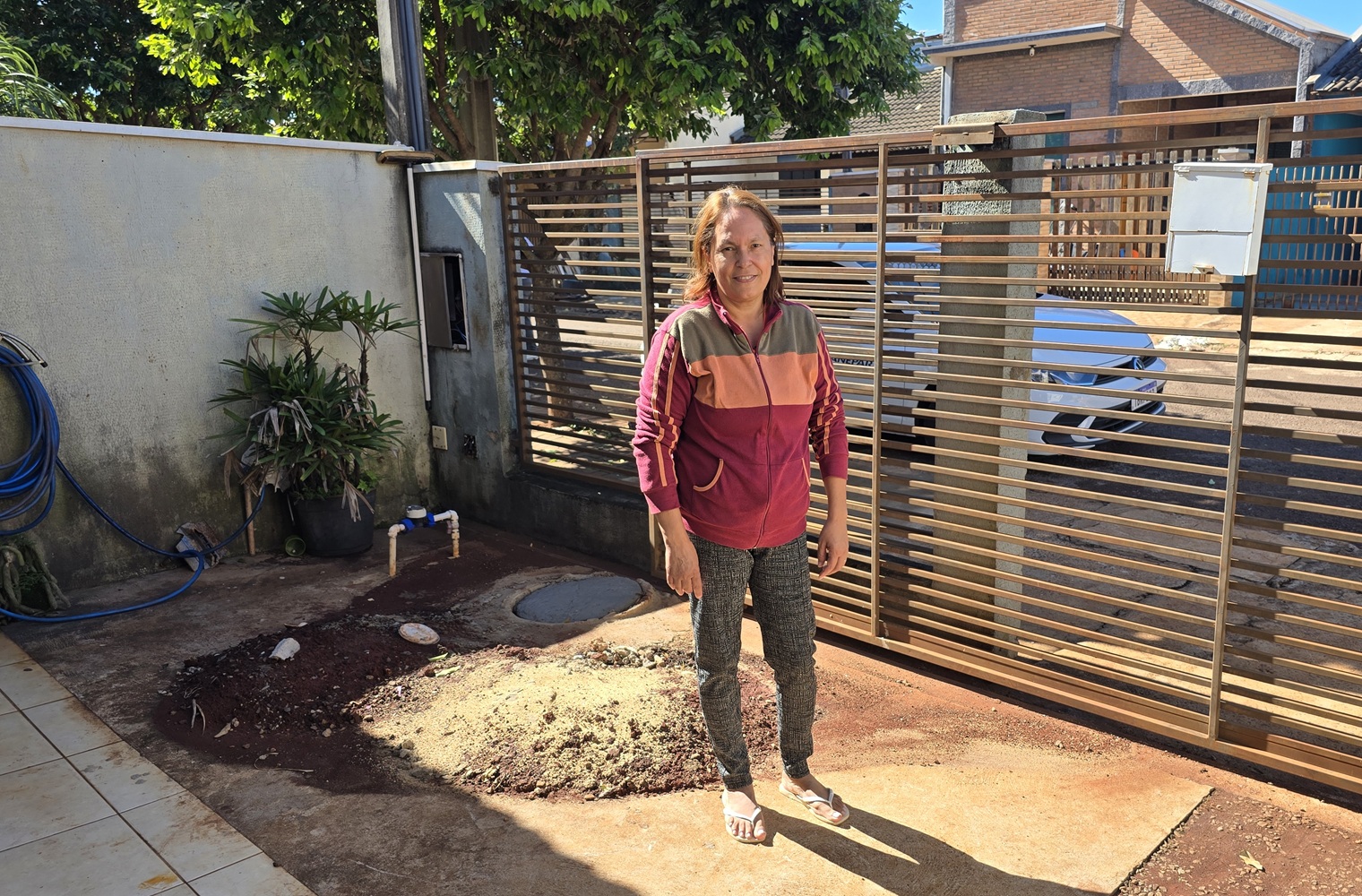 Mulher de casaco colorido e calça escura posa em pé no quintal de sua residência no Jardim Planalto. Ela está ao lado de uma tampa de inspeção de esgoto no chão, com terra onde a fossa foi aterrada. Ela está em frente a um portão de barras horizontais