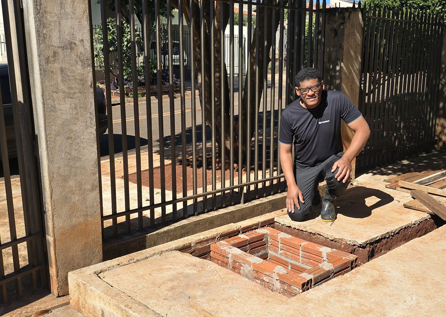 Morador sorri ajoelhado na calçada ao lado de uma caixa de inspeção ou passagem de esgoto recém-construída em alvenaria de tijolos. Ele está orgulhoso, pois foi ele quem fez a ligação de esgoto da sua casa. Ao fundo, uma grade metálica preta delimita o espaço residencial