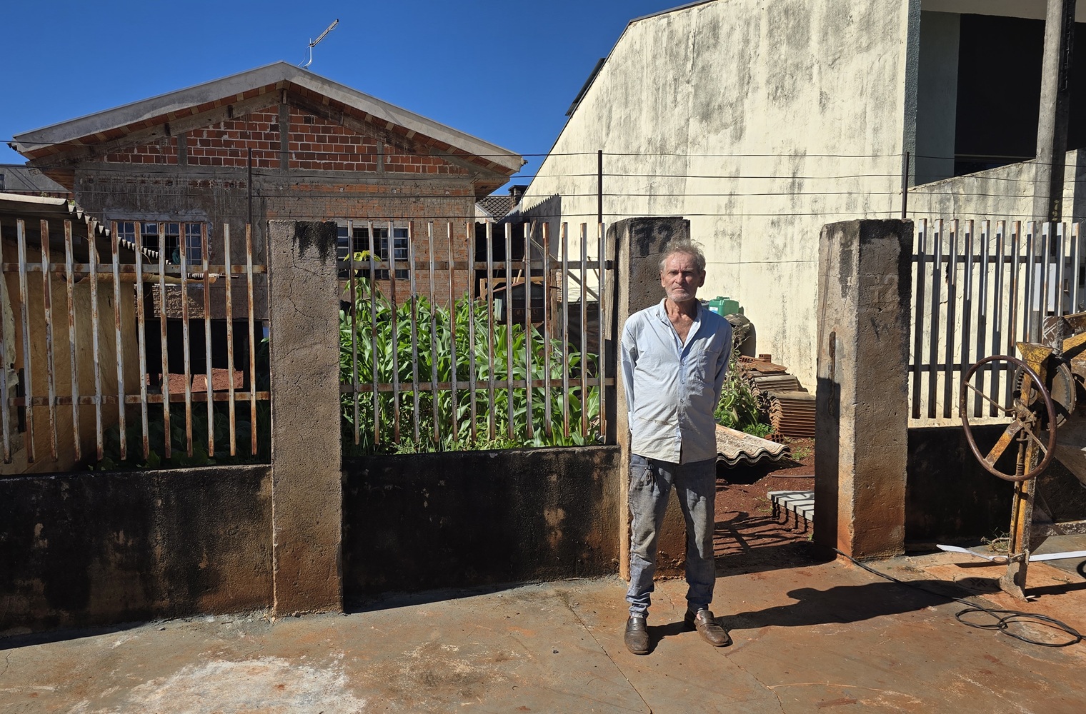 Homem de camisa clara e calça jeans posa em pé na calçada em frente ao portão de uma residência no Jardim Delgado. Ao fundo, aparecem casas de alvenaria sob um céu azul limpo