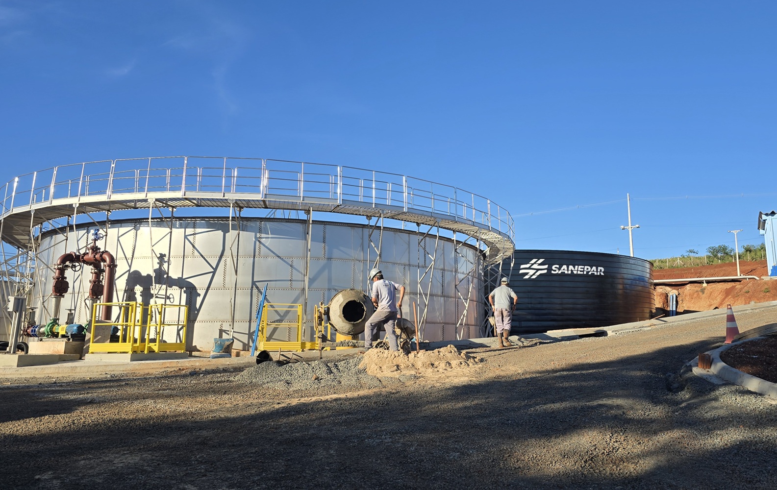 Dois operários trabalham no canteiro de obras de uma estação de tratamento de esgoto. Um deles opera uma betoneira móvel perto de um grande tanque metálico prateado com passarela superior. Ao fundo, um tanque preto exibe o logotipo da Sanepar sob o céu azul