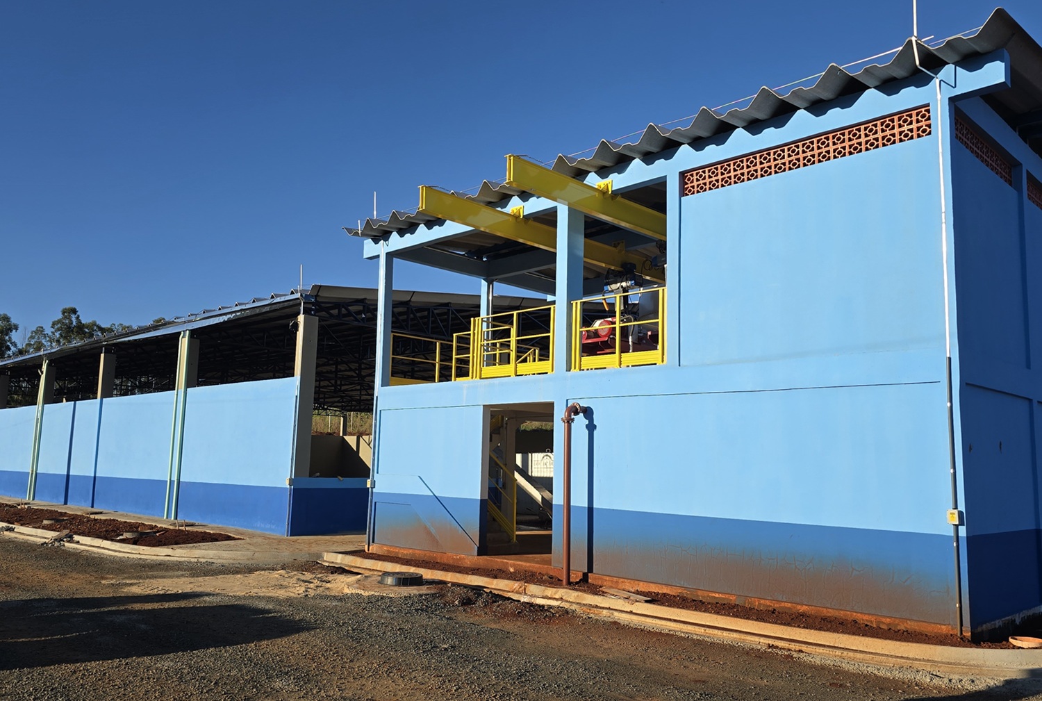 Prédio operacional com pé direito bem alto com paredes pintadas em tons de azul claro e escuro em uma estação de tratamento de esgoto. Anexo uma estrutura possui uma área aberta no piso superior com guarda-corpos amarelos e vigas metálicas, onde estão equipamentos do tipo centrífugas