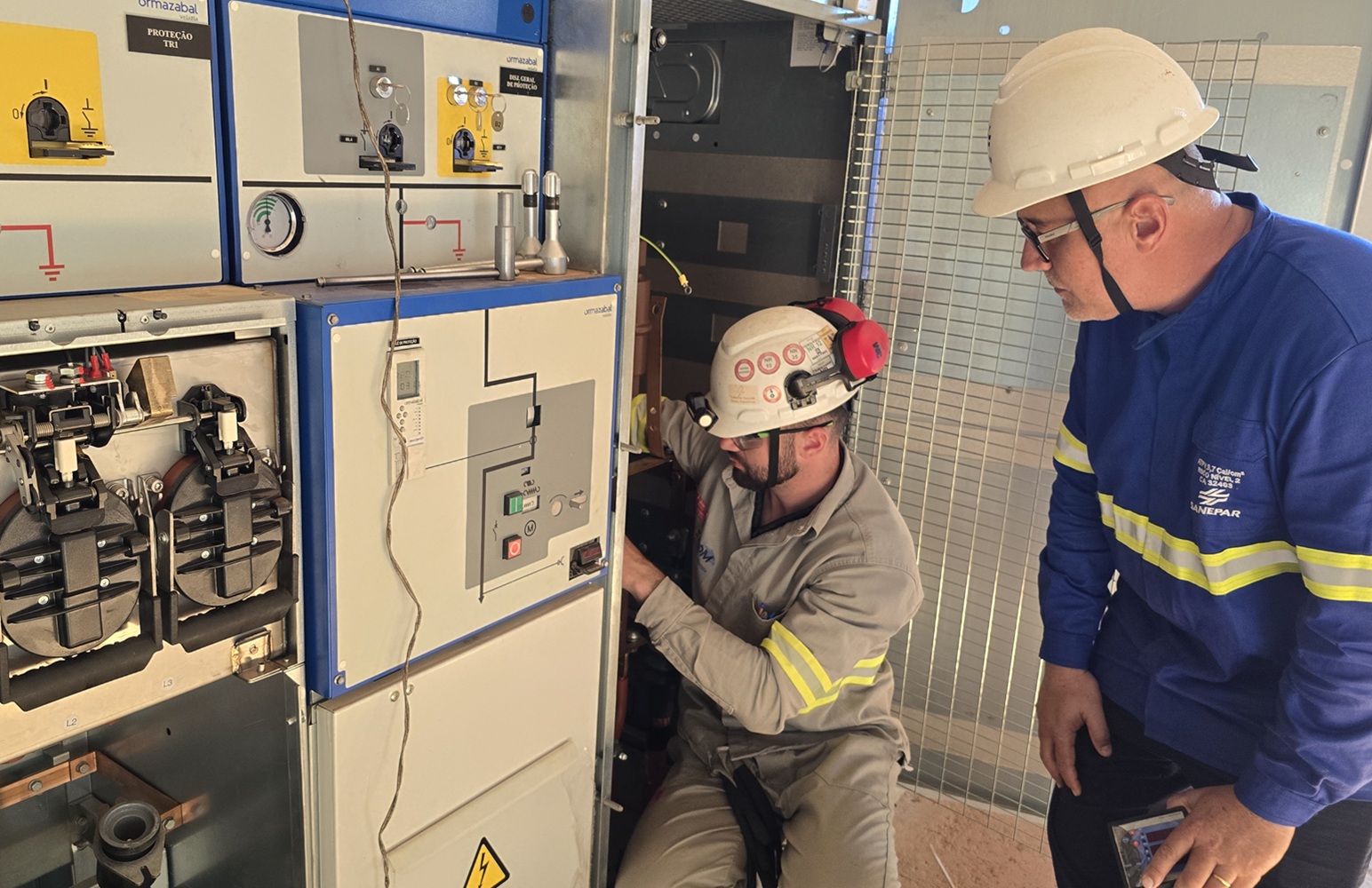 Dois técnicos trabalham em um painel elétrico. Um deles, agachado, realiza ajustes nos cabos internos utilizando capacete com lanterna e protetor auricular, sob a supervisão do colega de uniforme azul