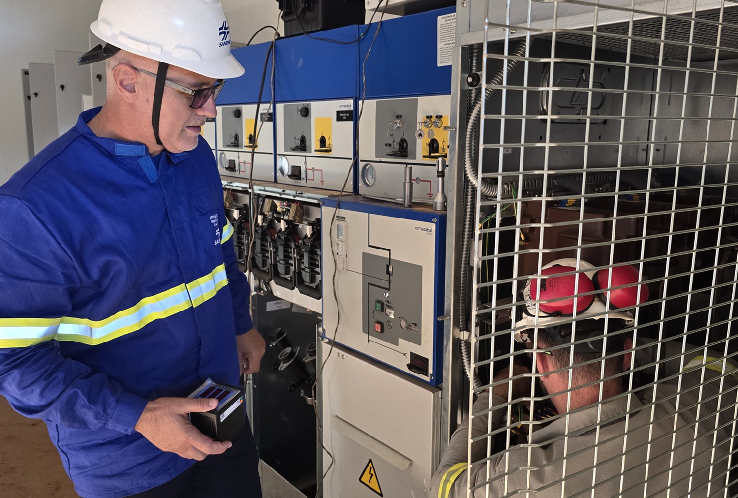 Técnico da Sanepar com uniforme azul e capacete branco observa outro profissional que realiza manutenção interna em um painel elétrico de alta tensão protegido por grade metálica
