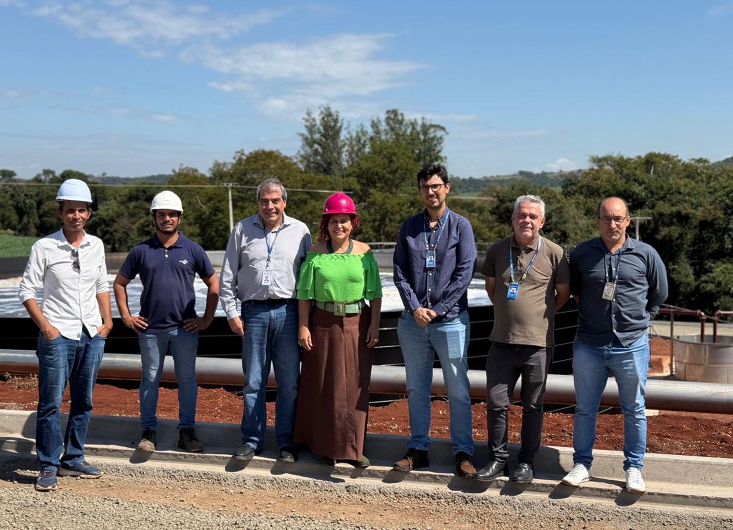Prefeita e membros da prefeitura, com equipe técnica da Sanepar posam para foto alinhados lado a lado no canteiro de obras da estação de tratamento de esgoto. A prefeita, ao centro, veste blusa verde e capacete rosa, enquanto os demais profissionais usam crachás, camisas casuais e capacetes brancos de proteção