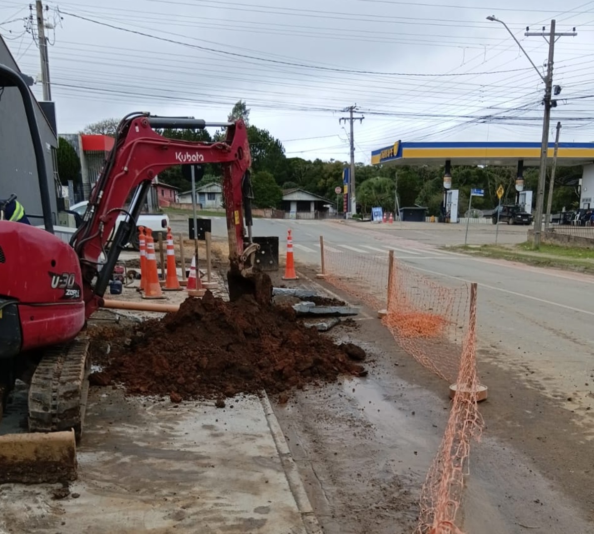 Mini escavadeira vermelha da marca Kubota realizando obras em uma calçada de rua. Há uma pilha de terra escura, cones de sinalização laranjas e uma rede de proteção laranja isolando a área. Ao fundo, vê-se um posto de combustíveis sob um céu nublado.