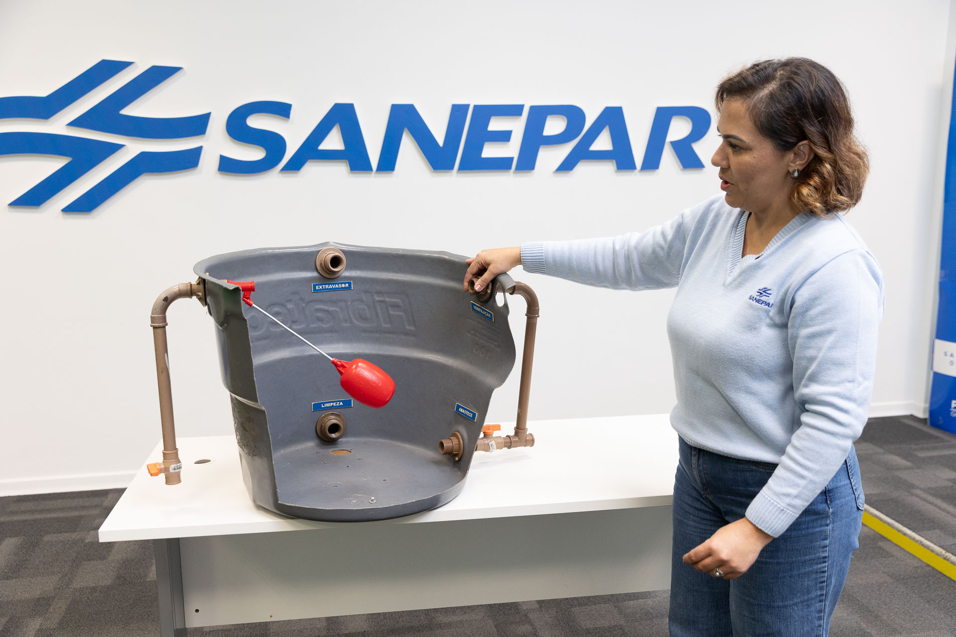 Mulher demonstra os componentes internos de uma caixa d'água utilizando uma maquete em corte que expõe a boia e as tubulações de entrada e saída. Ao fundo, parede branca com o logotipo azul da Sanepar.