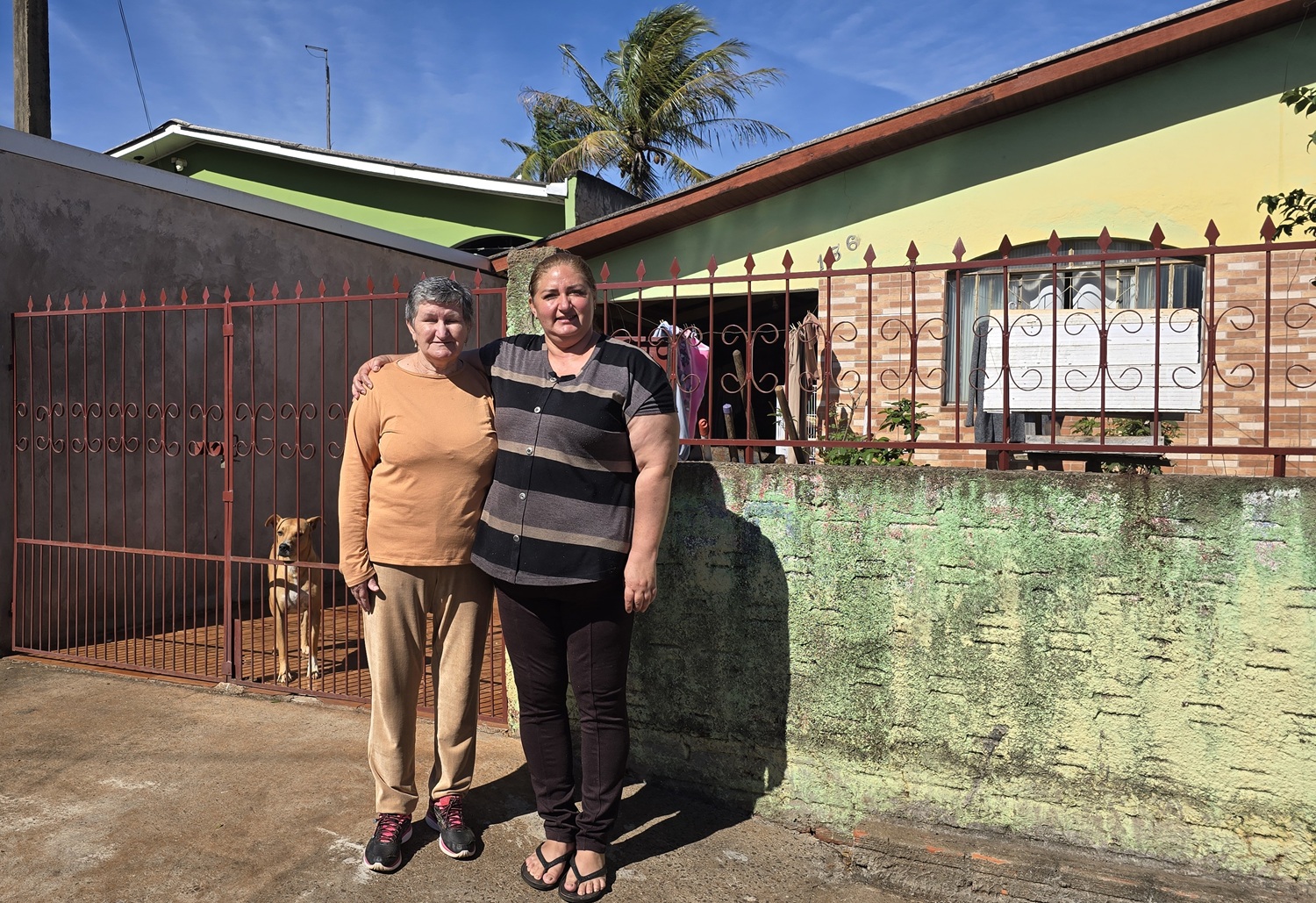 mãe e filha abraçadas na frente de casa, uma senhor com cabelos claros e curtos e a filha sorrindo. no fundo a cachorra caramelo aparece no portão
