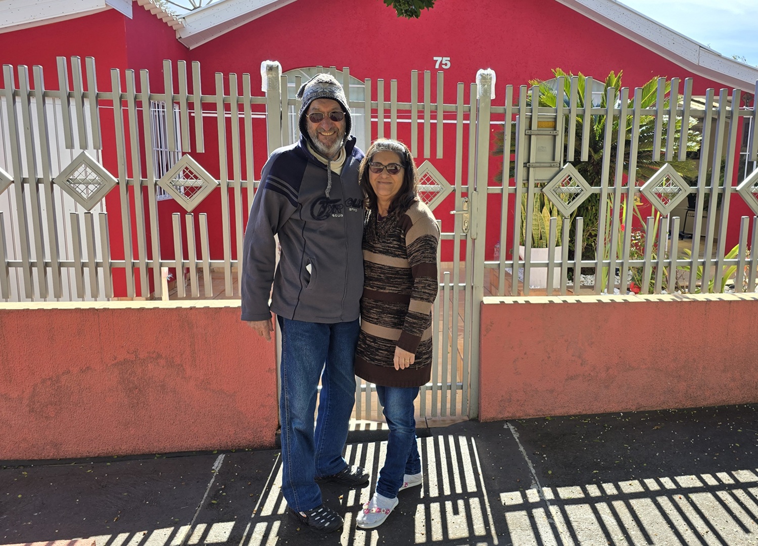casal em frente ao seu imóvel, ele alto e vestido roupas de lã, incluindo um gorro, ela bem mais baixa abraçada