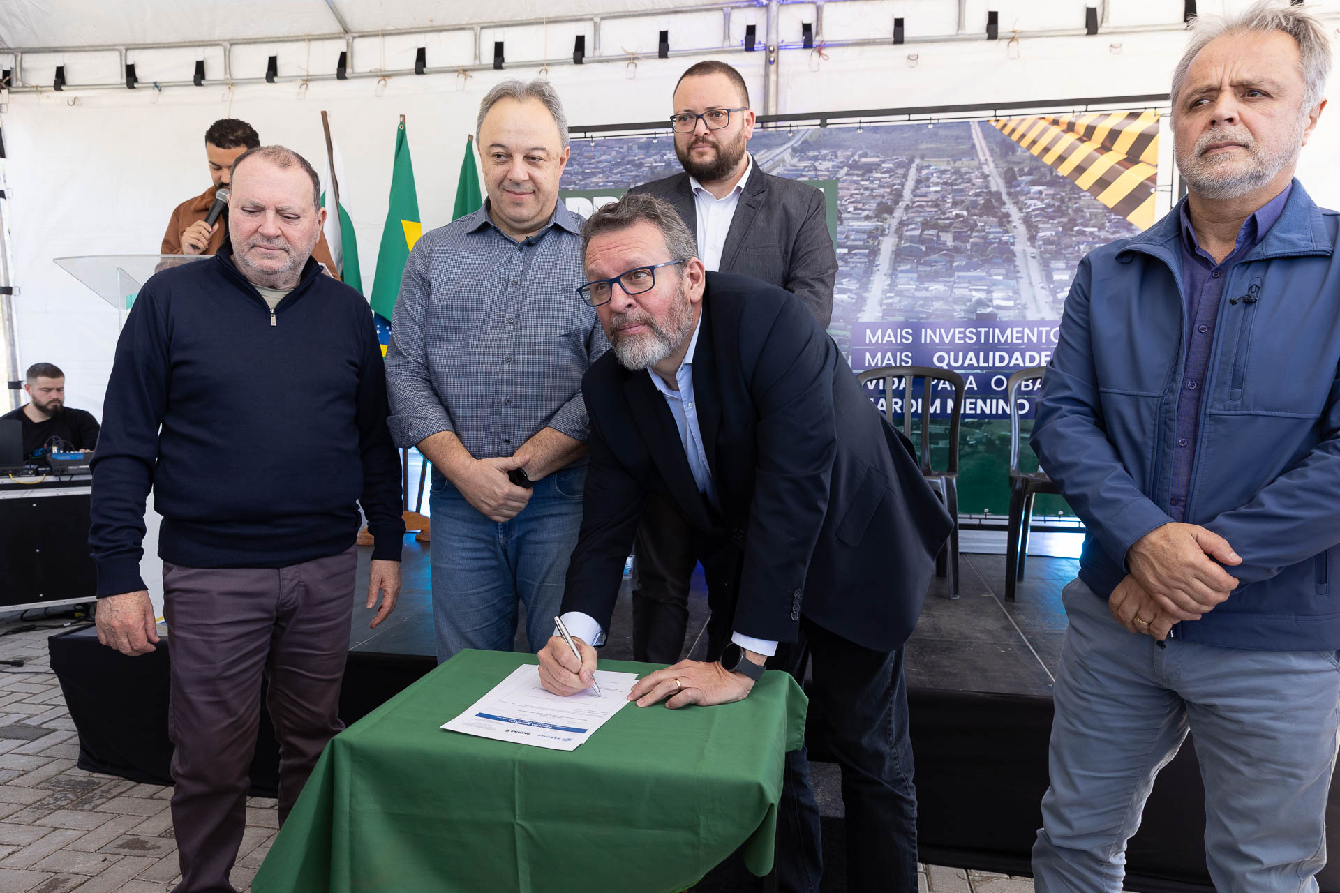 Homem de óculos e terno escuro inclina-se para assinar um documento sobre uma mesa com toalha verde. Ao seu redor, outros quatro homens acompanham o ato em um palco sob uma tenda. Ao fundo, há bandeiras e um banner com a imagem aérea de um bairro.