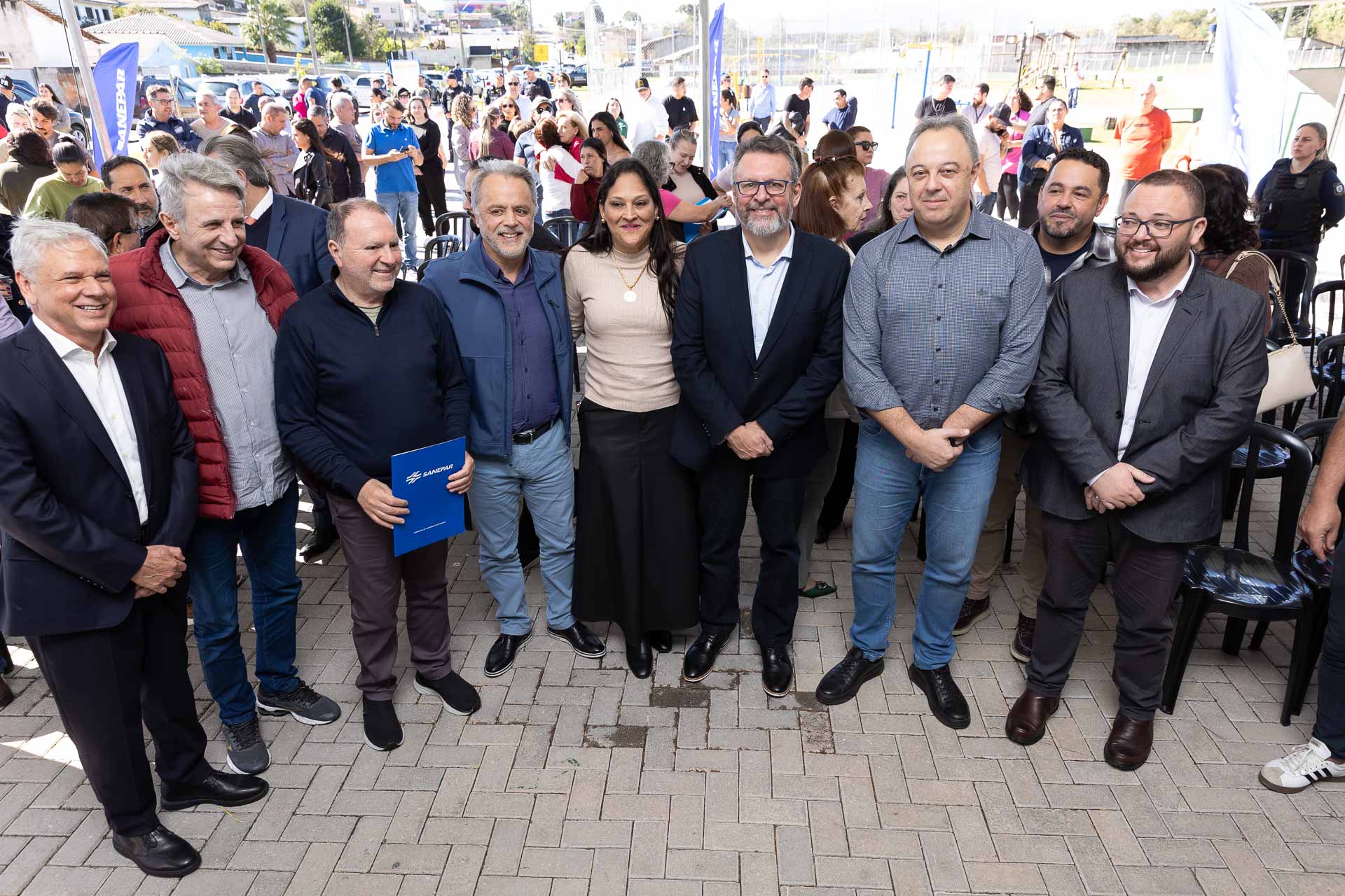 Grupo de autoridades e representantes da Sanepar posa sorrindo para foto em um evento público ao ar livre. Um dos homens segura uma pasta azul com o logotipo da Sanepar, enquanto uma grande quantidade de pessoas acompanha o evento ao fundo.