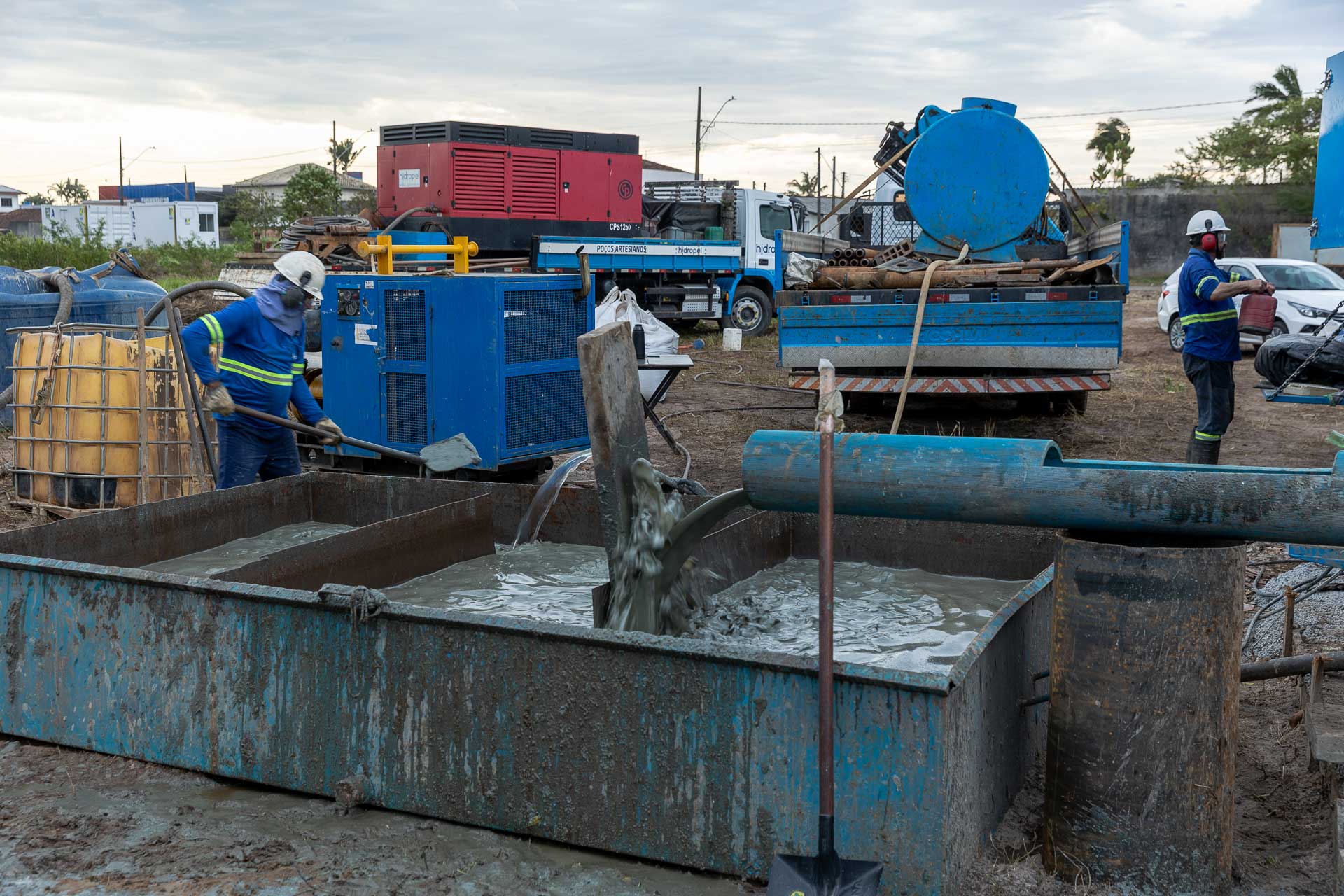 Tubulação despejando fluido cinzento de perfuração em um tanque de contenção de metal, com trabalhadores da equipe técnica operando ao redor.