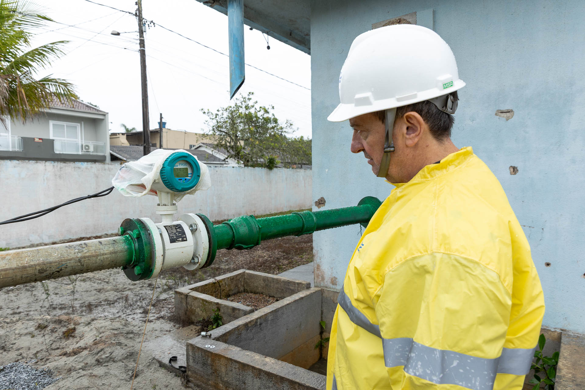 Técnico com capacete branco e capa de chuva amarela observa atentamente o visor de um medidor de vazão digital acoplado a uma tubulação verde.