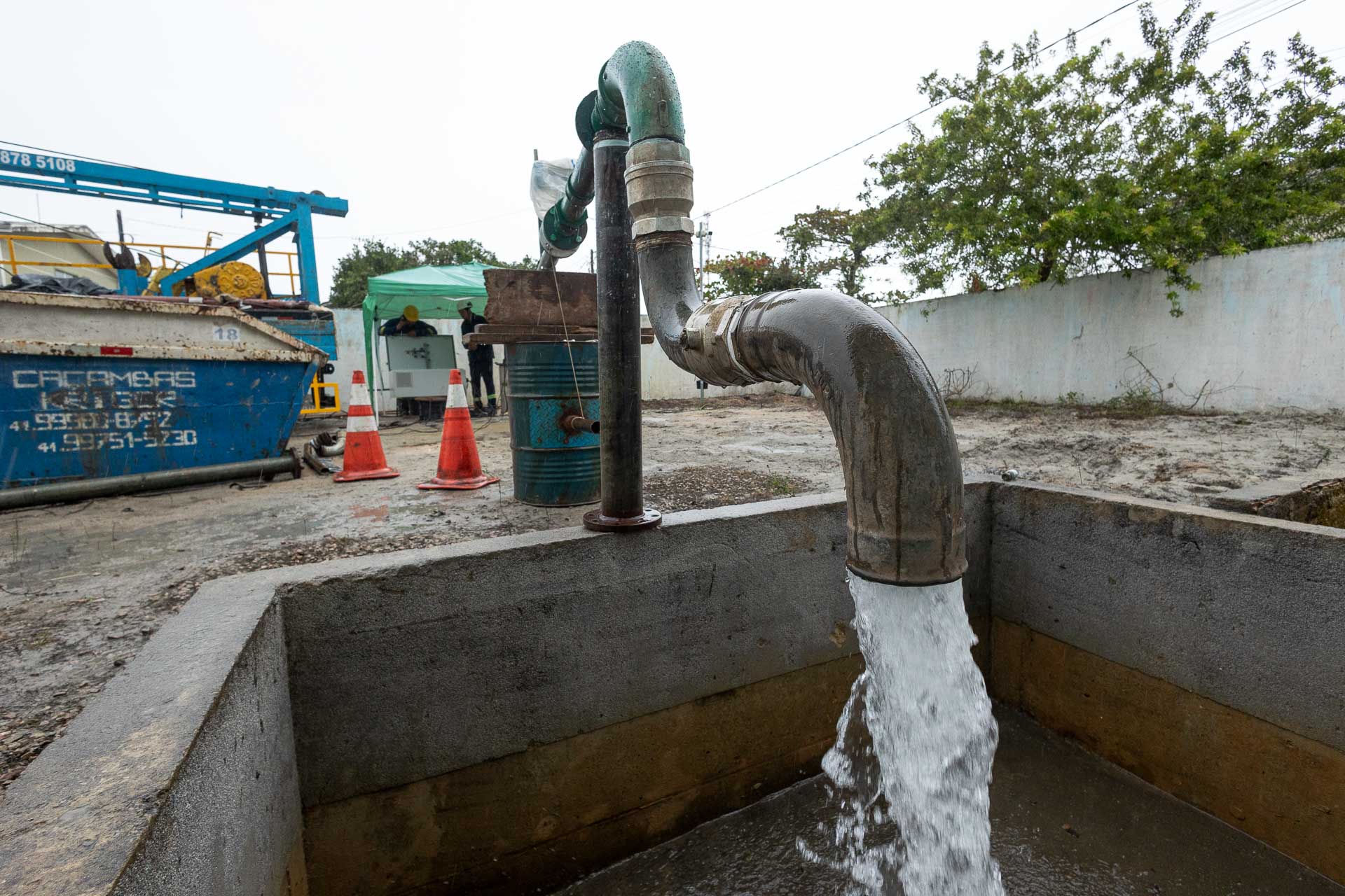 Água jorrando com abundância de uma tubulação metálica para dentro de um tanque de concreto. Ao fundo, dois trabalhadores com capacetes operam equipamentos de perfuração em um canteiro de obras.