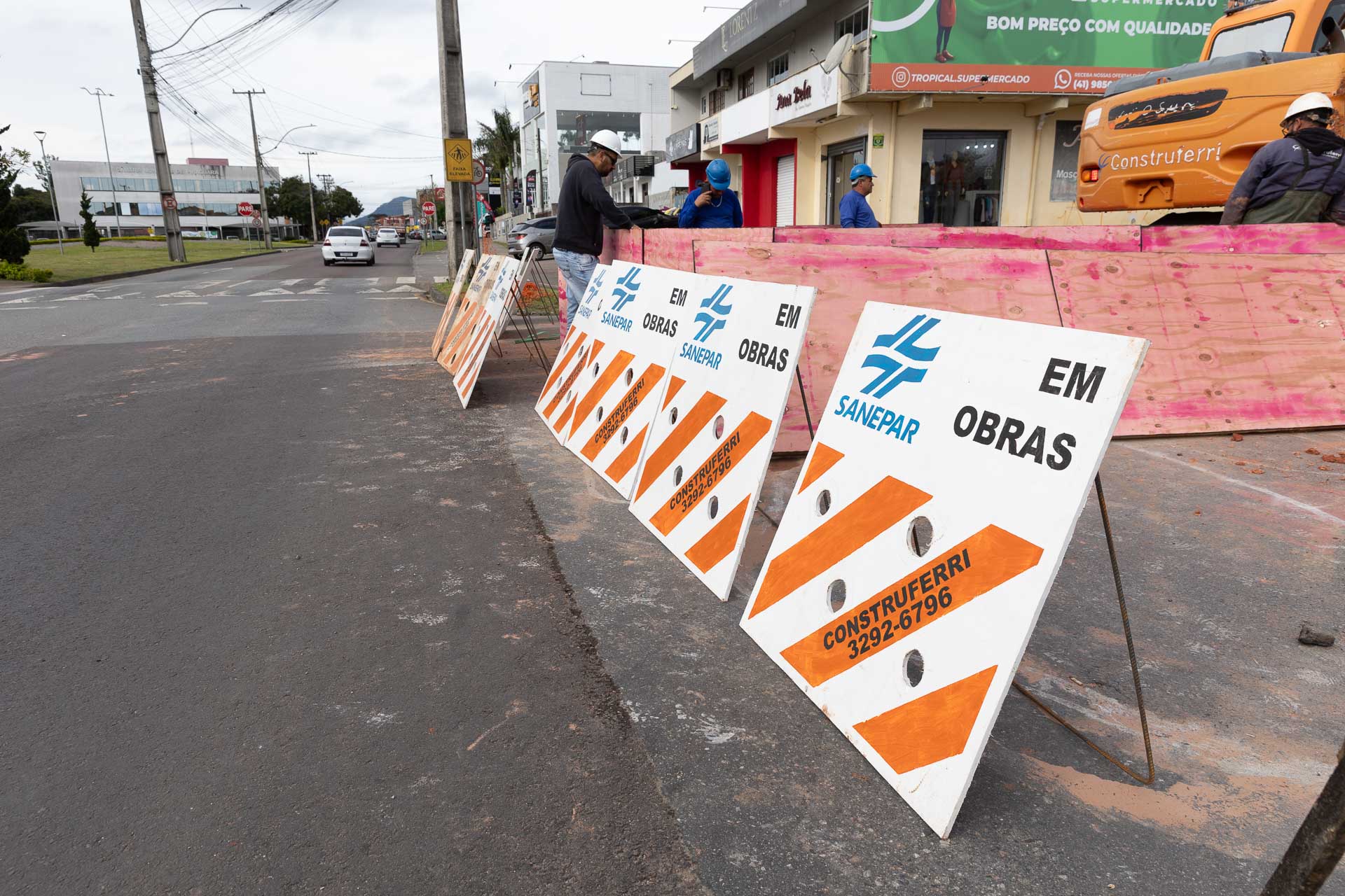 Cavaletes de sinalização com listras brancas e laranjas, logotipo da Sanepar e o texto "EM OBRAS", bloqueiam parte da via, isolando a área onde trabalhadores e maquinário atuam ao fundo.