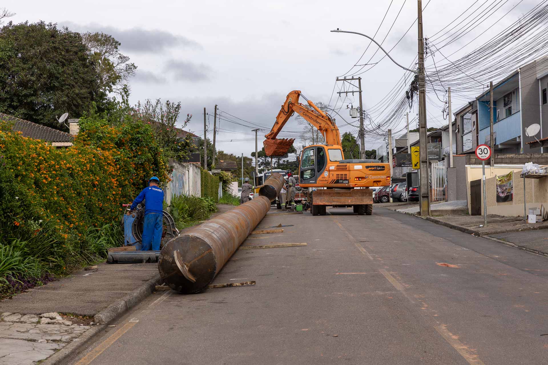 Escavadeira laranja ergue um longo tubo metálico no meio de uma rua residencial, com operários de uniforme azul acompanhando a movimentação da estrutura.