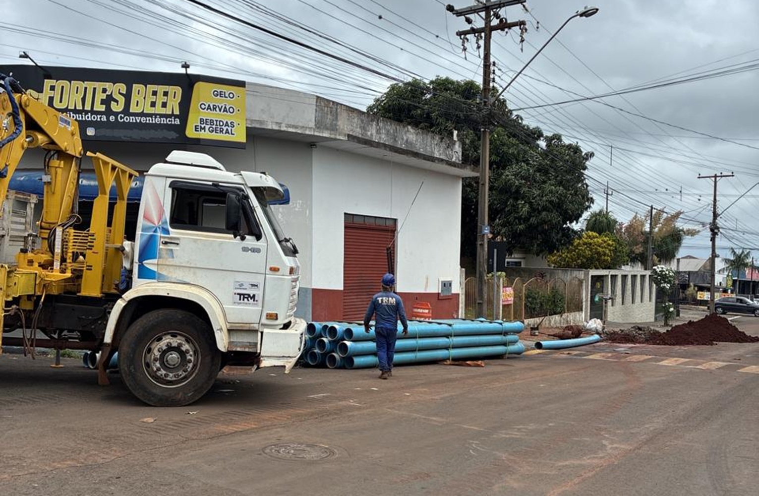 numa rua, um caminhão aparece no canto da foto, no chão ao lado, tem uma pilha de tubulações de água em PVC na cor azul e tem também um trabalhador da obra vestido de azul