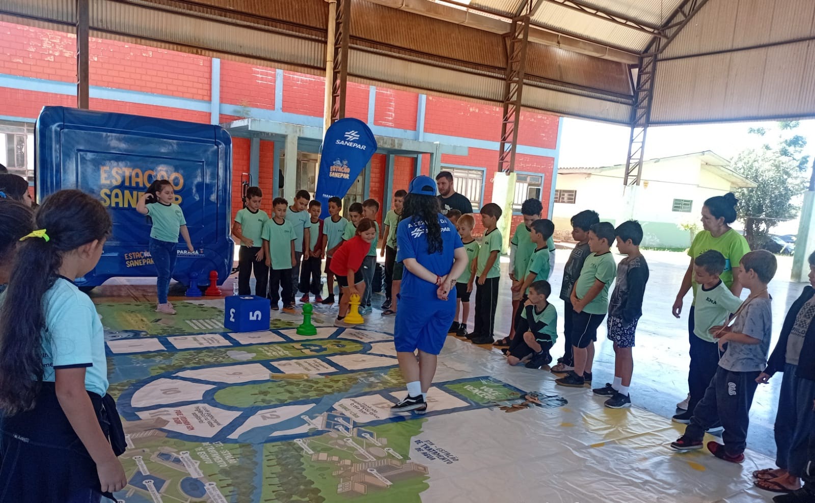 Alunos em fila participando de um jogo de tabuleiro gigante e interativo no chão, mediado por monitores da Sanepar em uma quadra coberta escolar.