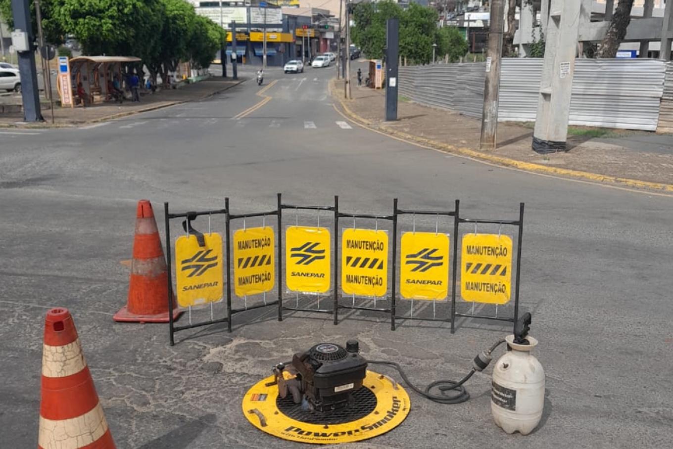 Equipamento de aplicação de fumaça posicionado sobre o poço de visita na rua, isolado por gradis amarelados de sinalização da Sanepar com a palavra "Manutenção" e cones de trânsito