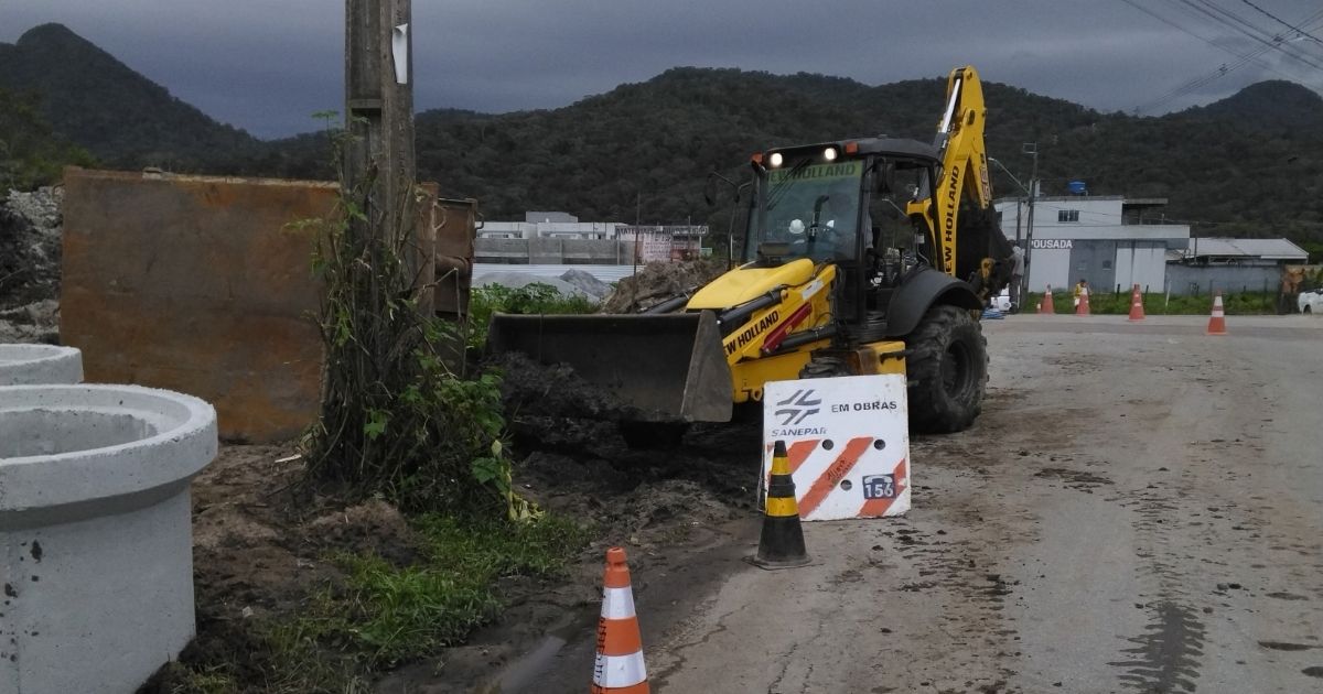 Retroescavadeira amarela movimenta terra na lateral de uma rua asfaltada, próxima a peças circulares de concreto. Cones de trânsito laranjas e uma placa da Sanepar com o aviso "Em obras" e o telefone 156 sinalizam o local.