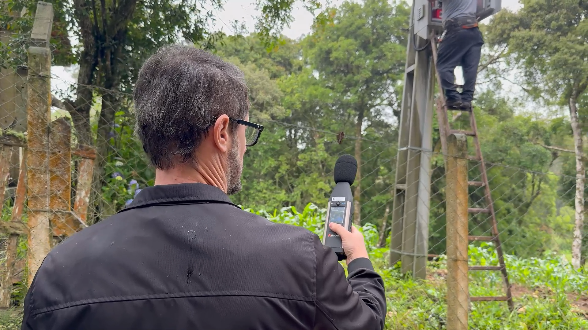 Fotografia em plano médio focada nas costas de um técnico da Sanepar, um homem de cabelos grisalhos e óculos, que segura um decibelímetro digital apontado para cima para medir a intensidade do som. Ao fundo, após uma cerca de arame, outro operador trabalha subido em uma escada de madeira apoiada contra uma estrutura de concreto alta, realizando ajustes em uma caixa de comandos eletrônicos. O ambiente ao redor é repleto de mata verde densa.