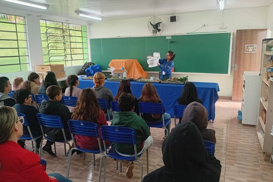 Educadora da Sanepar em sala de aula segura maquete de vaso sanitário durante palestra de conscientização sobre saneamento para alunos