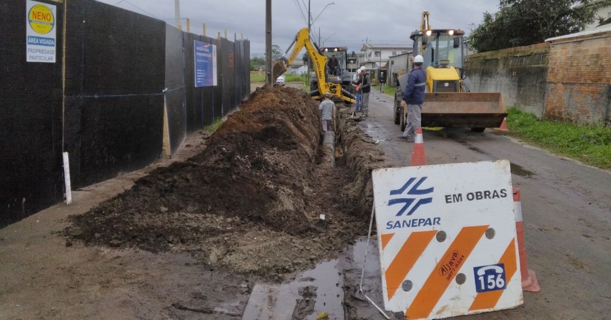 Vala extensa escavada na margem de uma rua asfaltada para a passagem de tubulação. Mais adiante, operários e retroescavadeiras dão andamento à obra. Em primeiro plano, há uma placa de sinalização da Sanepar com os dizeres "Em obras" e o telefone 156.