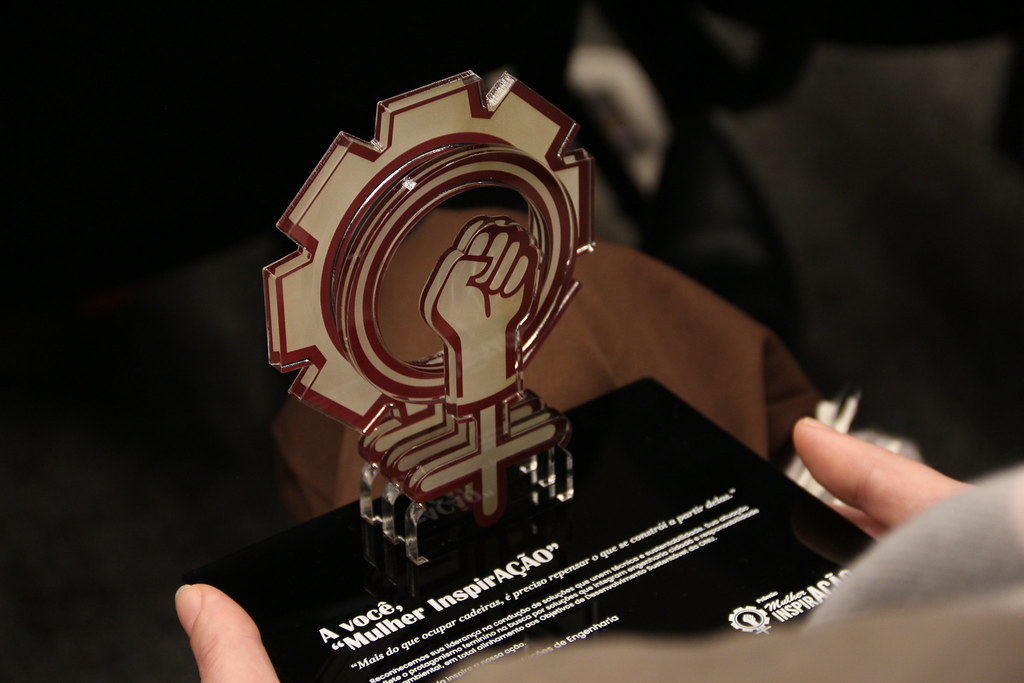 Troféu do Prêmio Mulher InovAÇÃO, em formato de engrenagem com um punho fechado ao centro. Ele é dourado e tem uma base quadrada preta.